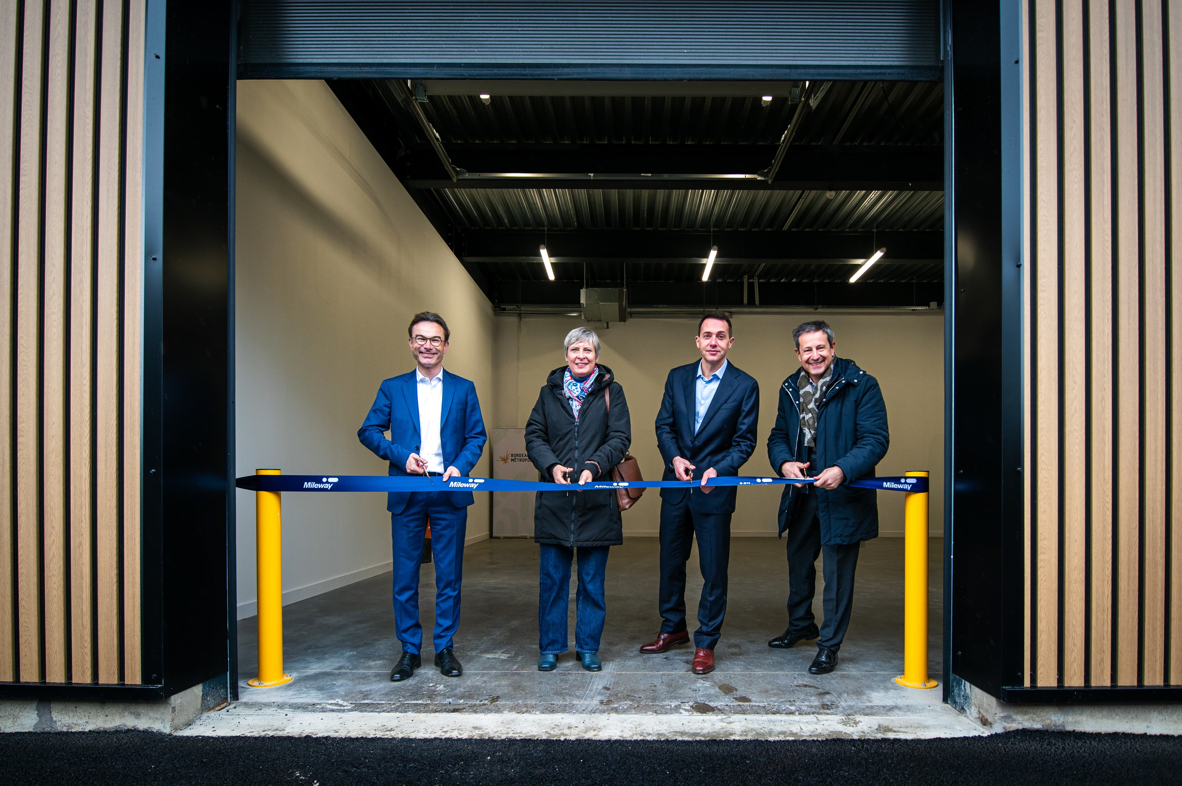 Four people in formal attire stand in front of a building, holding a ribbon for a ribbon-cutting ceremony at the entrance of an industrial facility.