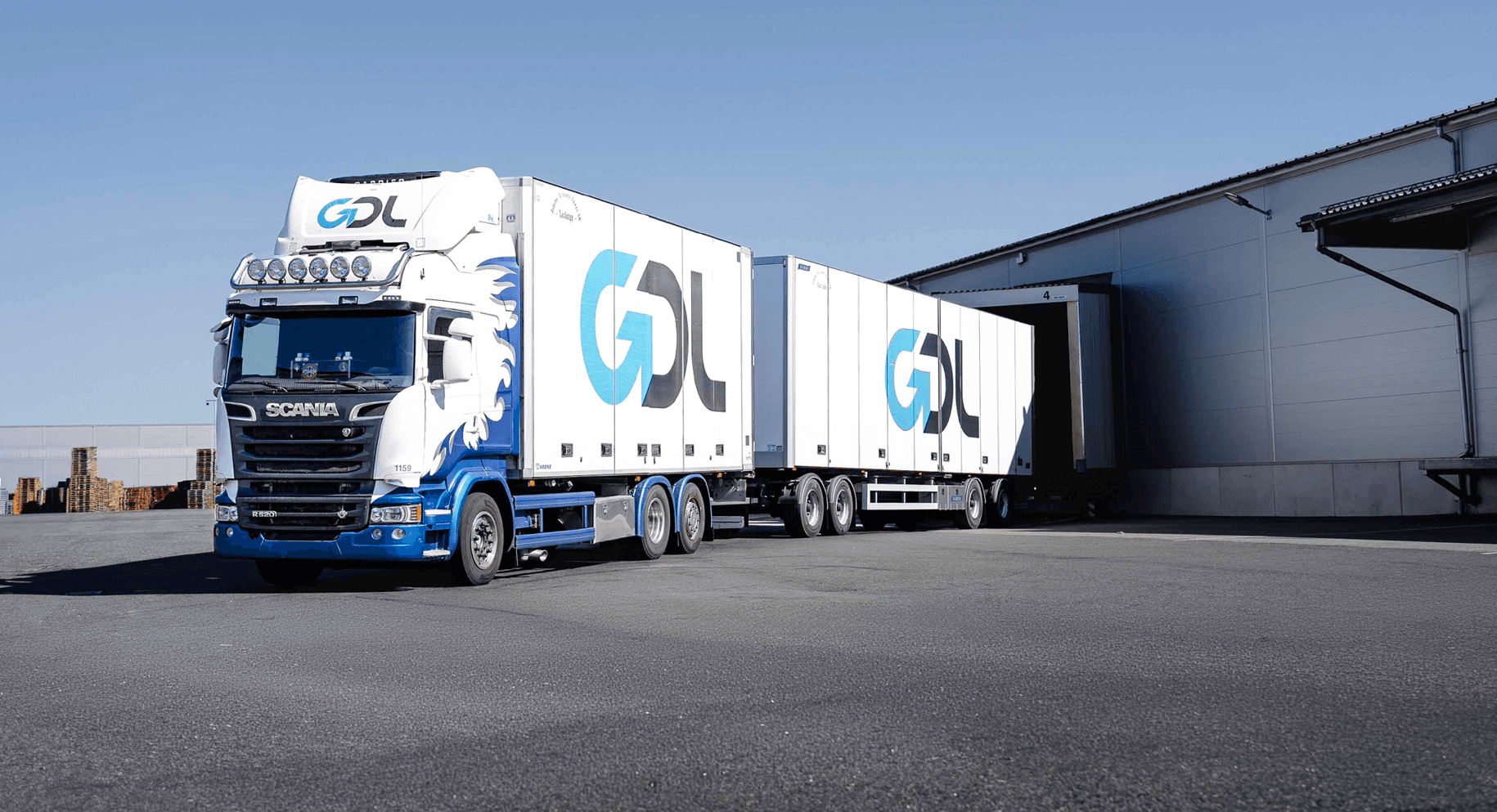 A white and blue Scania semi-truck with a GDL logo is parked outside a warehouse loading dock under a clear sky.