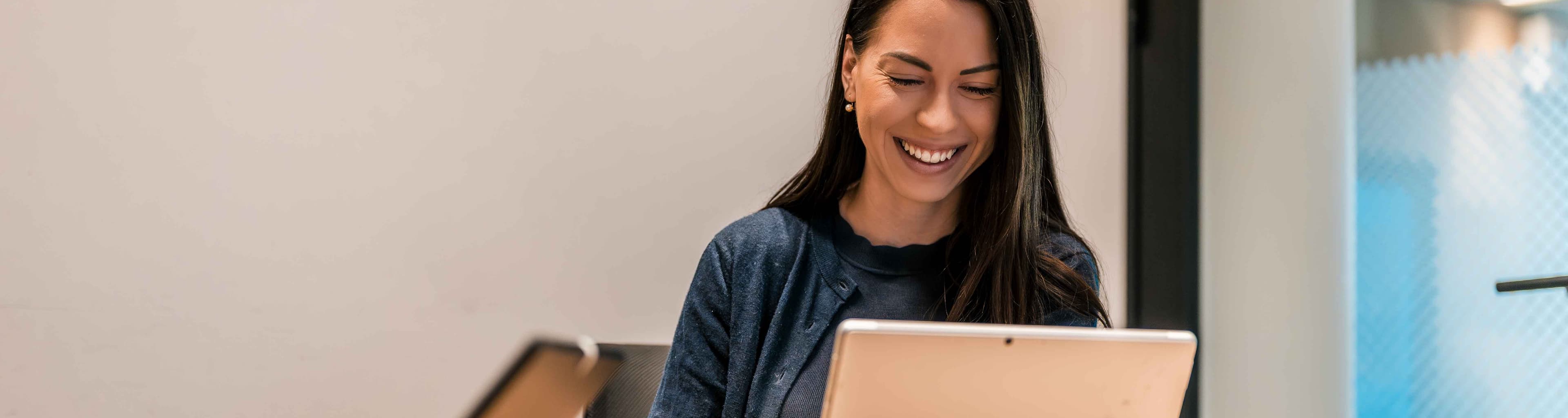 A woman sits at a desk using a Microsoft Surface device, smiling, with another person and tablet partially visible beside her in a modern office setting.