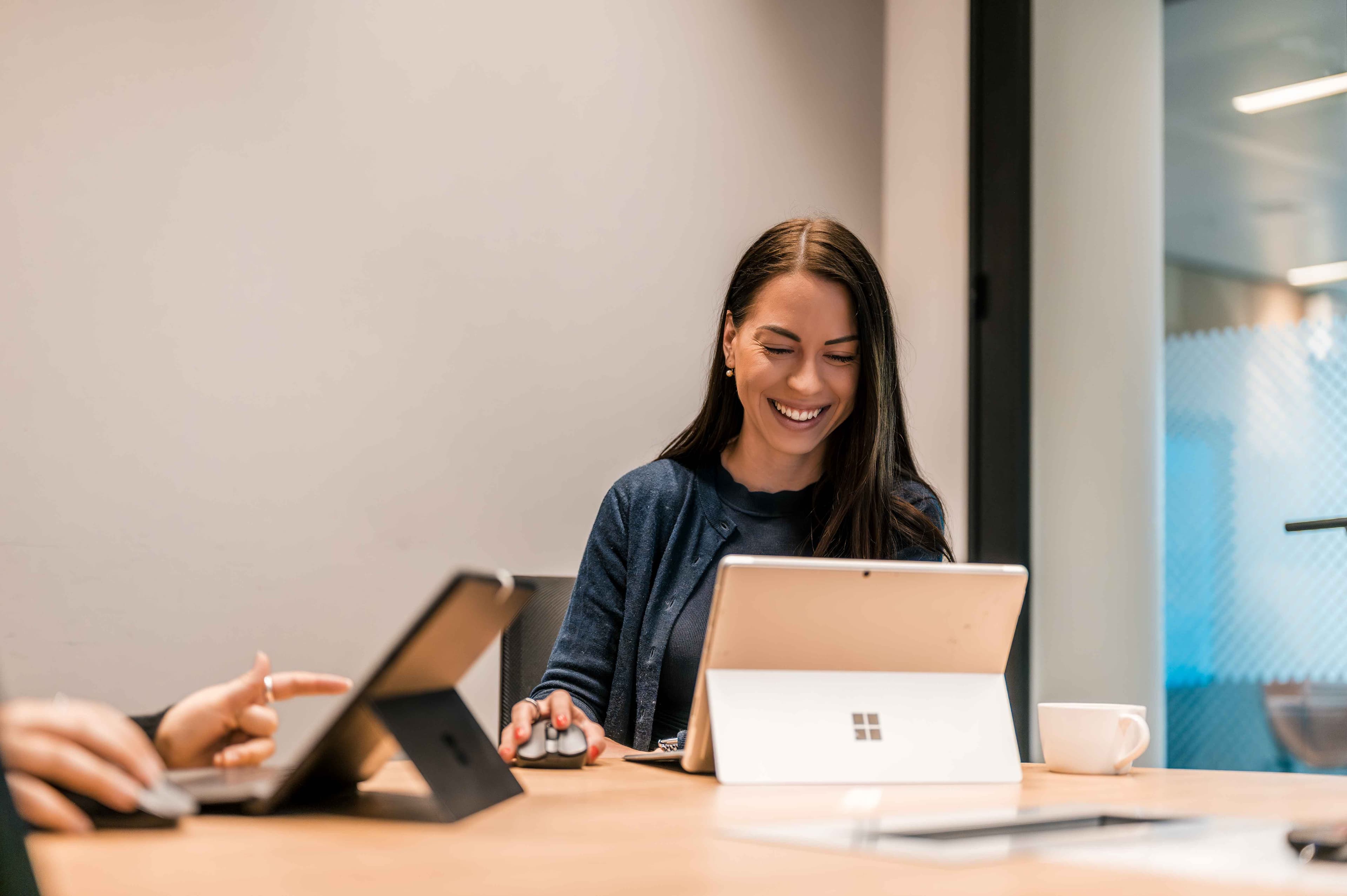 Una mujer está sentada en un escritorio utilizando un portátil Microsoft Surface, sonriendo, con otra persona trabajando en una tableta cerca en un entorno de oficina moderno.