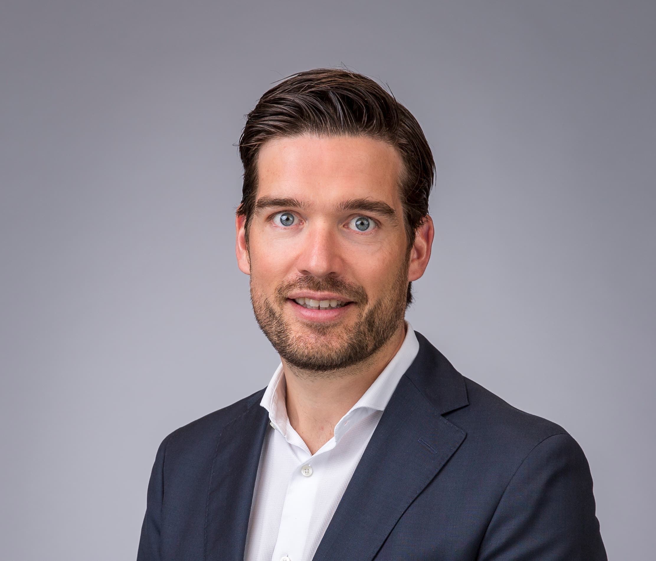 Een persoon in een donker pak en wit overhemd staat tegen een effen lichtgrijze achtergrond.