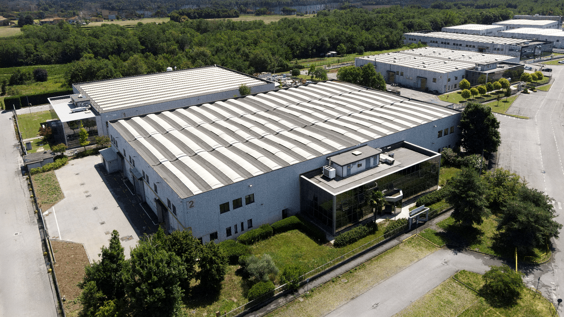 A large industrial warehouse with a curved metal roof, surrounded by greenery and similar buildings, seen from above.