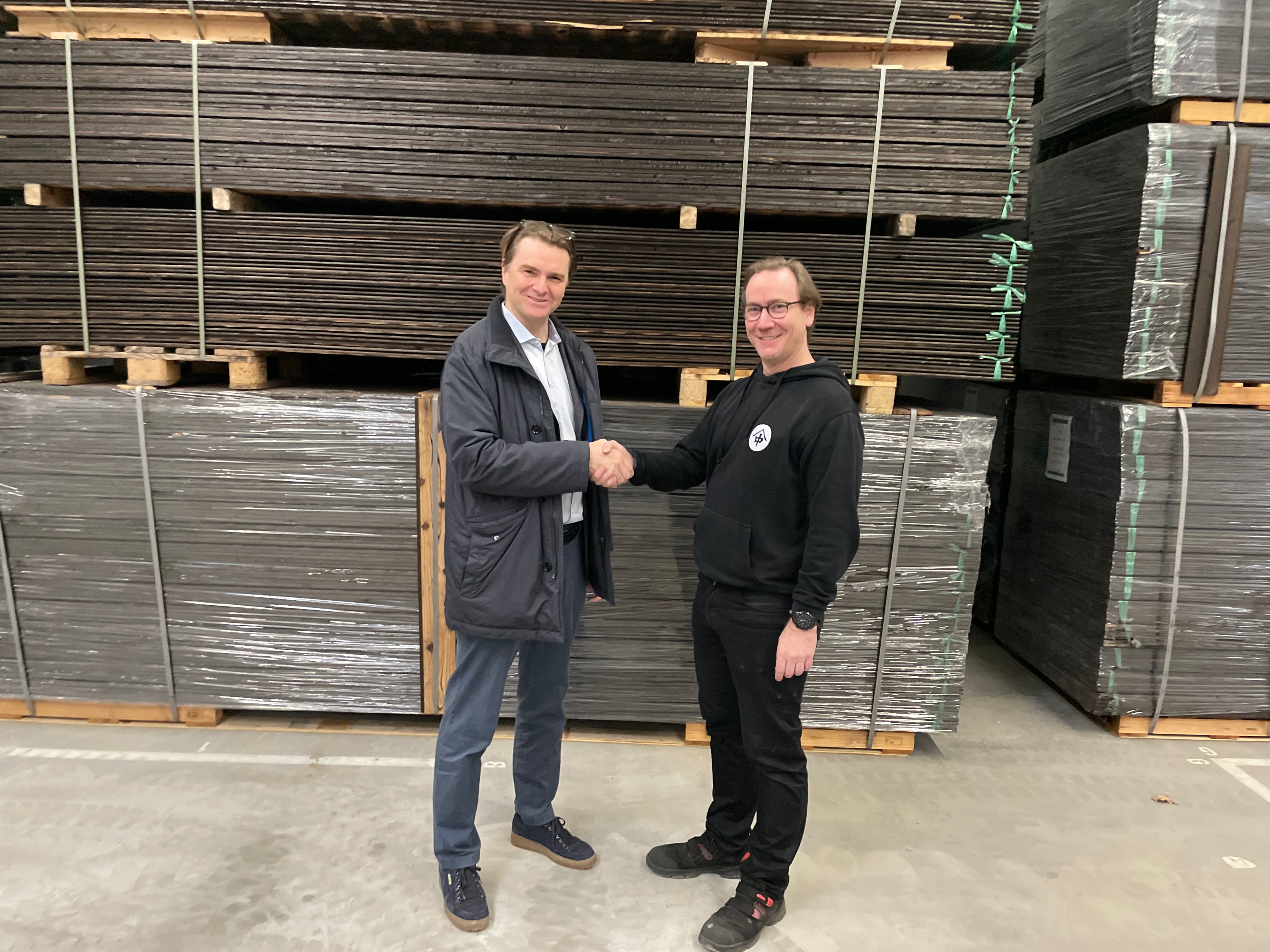 Two people shake hands in a warehouse, standing in front of stacked pallets of materials.