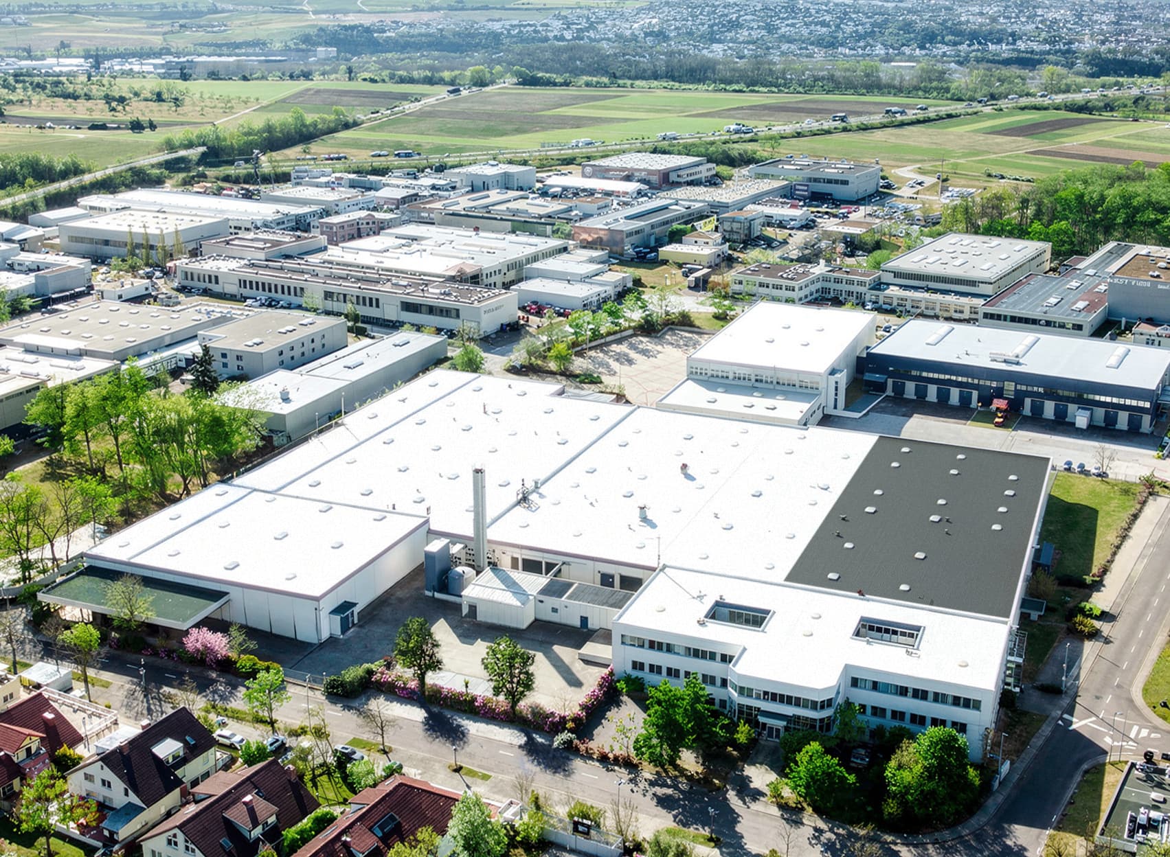 Aerial view of an industrial estate with large warehouses, nearby roads, greenery, and a residential area in the distance.