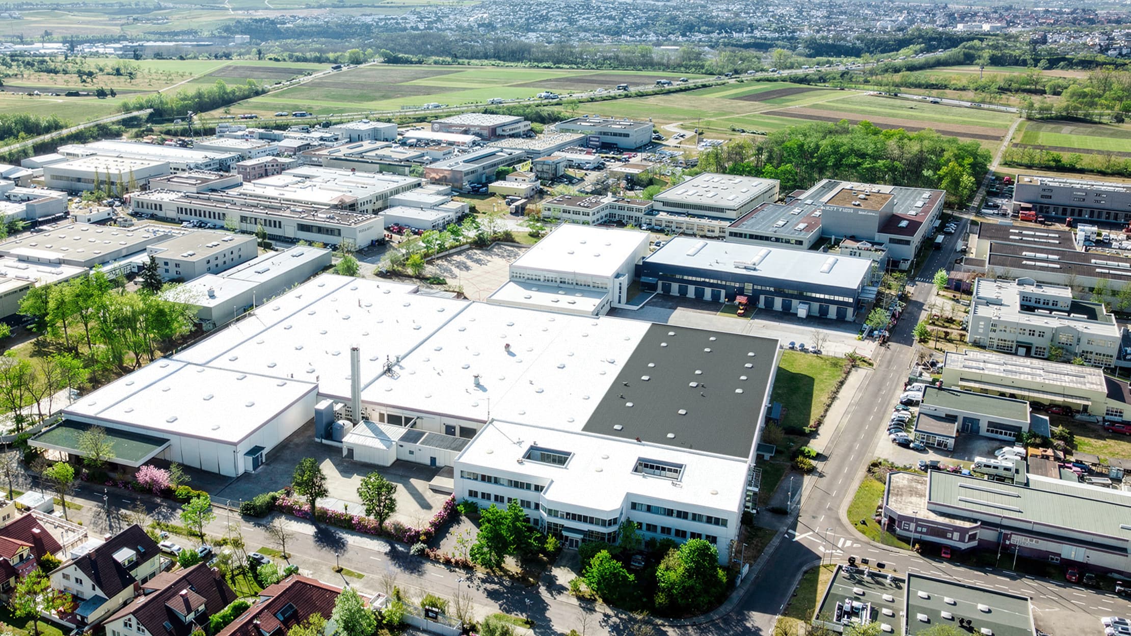 Luftaufnahme eines Industrieparks mit großen Lagerhallen, Parkplätzen und Grünflächen inmitten von Straßen und nahe gelegenen Wohngebieten.