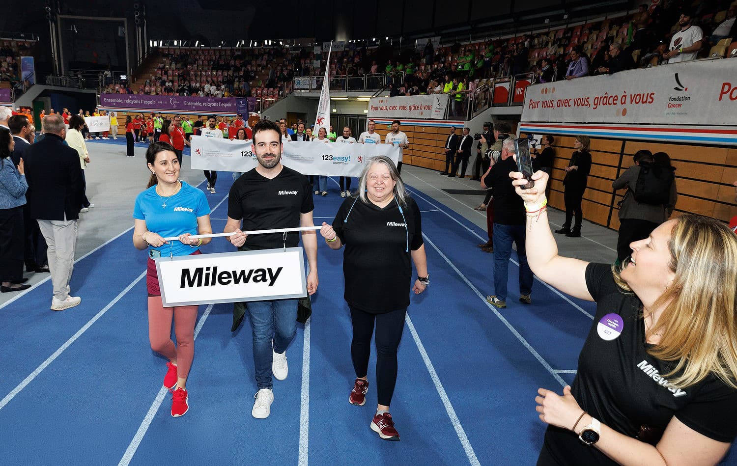 Four people in Mileway shirts walk on an indoor track holding a sign, with more groups and spectators visible in the background.