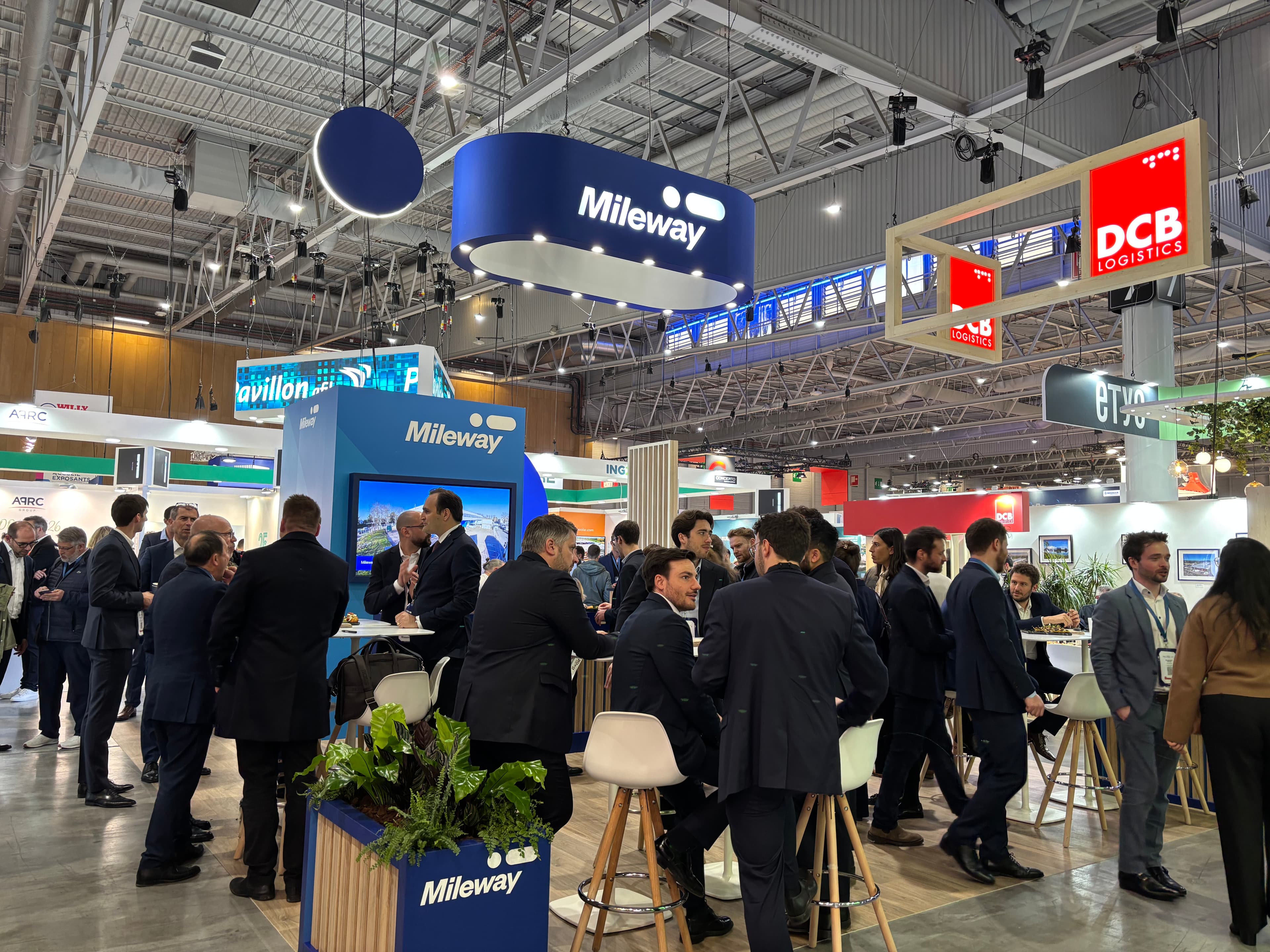People in business attire networking and conversing at a busy trade fair, with Mileway stand in the background.