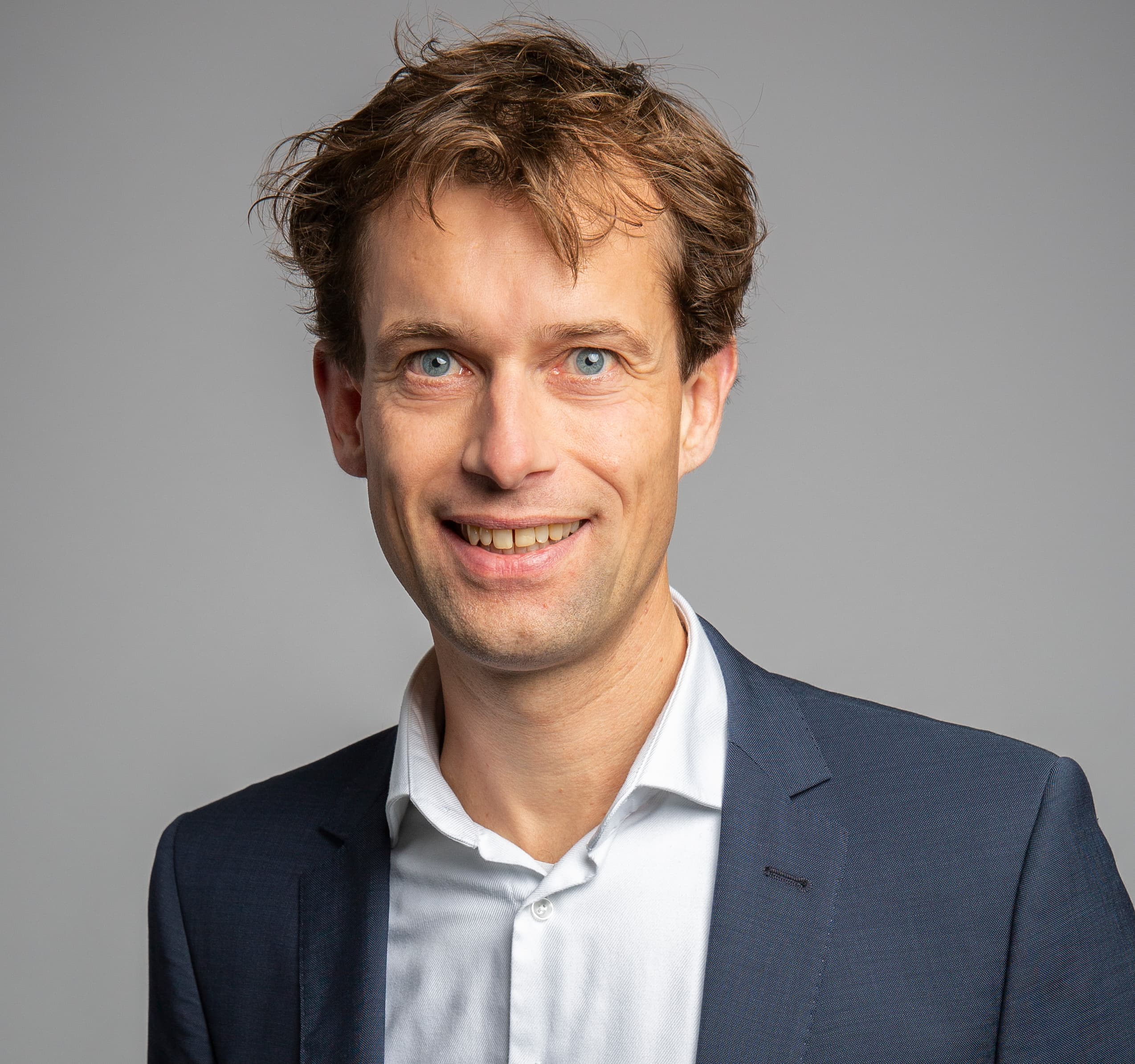 A person in a dark suit jacket over a white button-up shirt stands and smiles in front of a plain grey background.