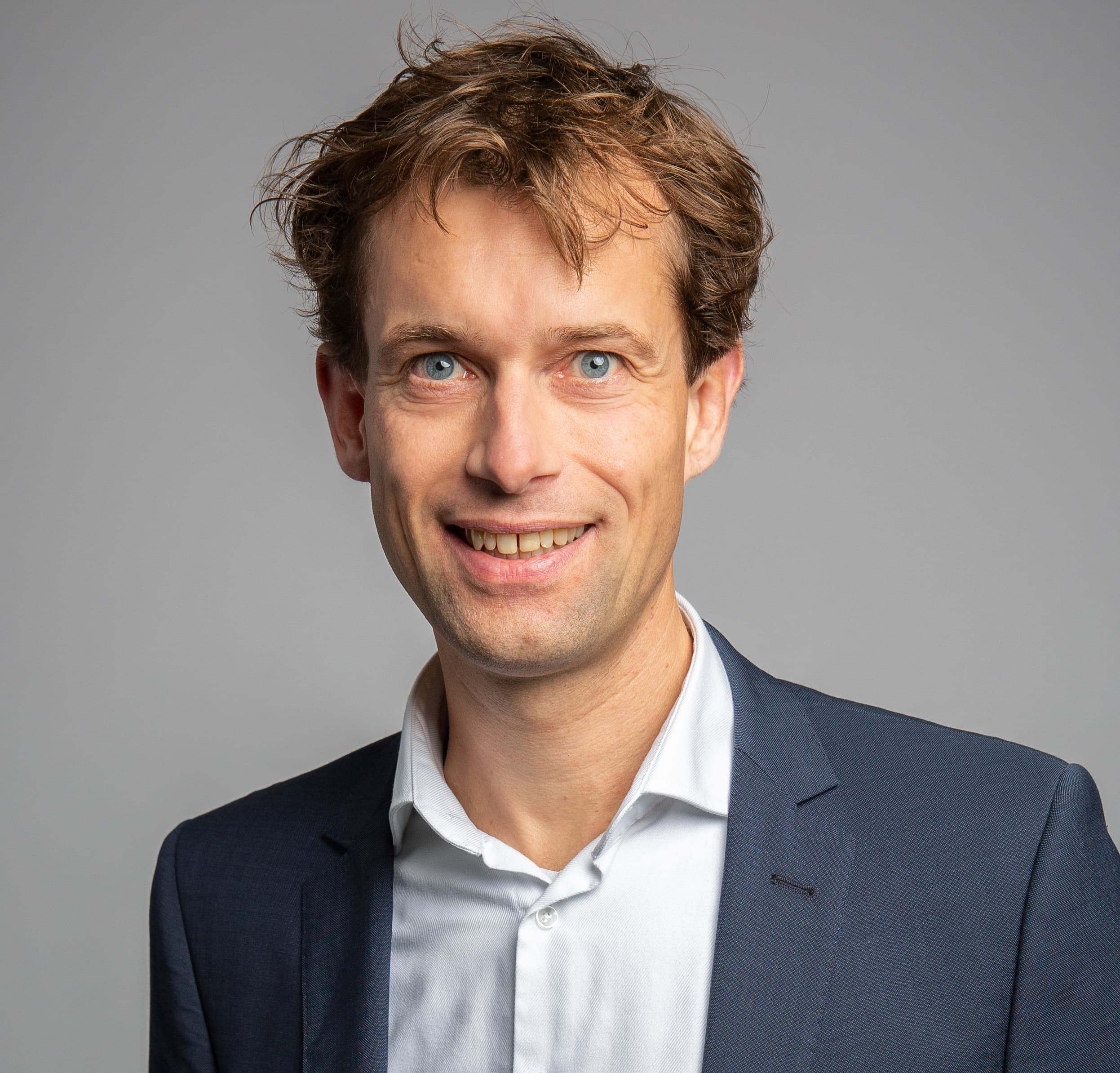 A person in a dark suit jacket over a white button-up shirt stands and smiles in front of a plain grey background.