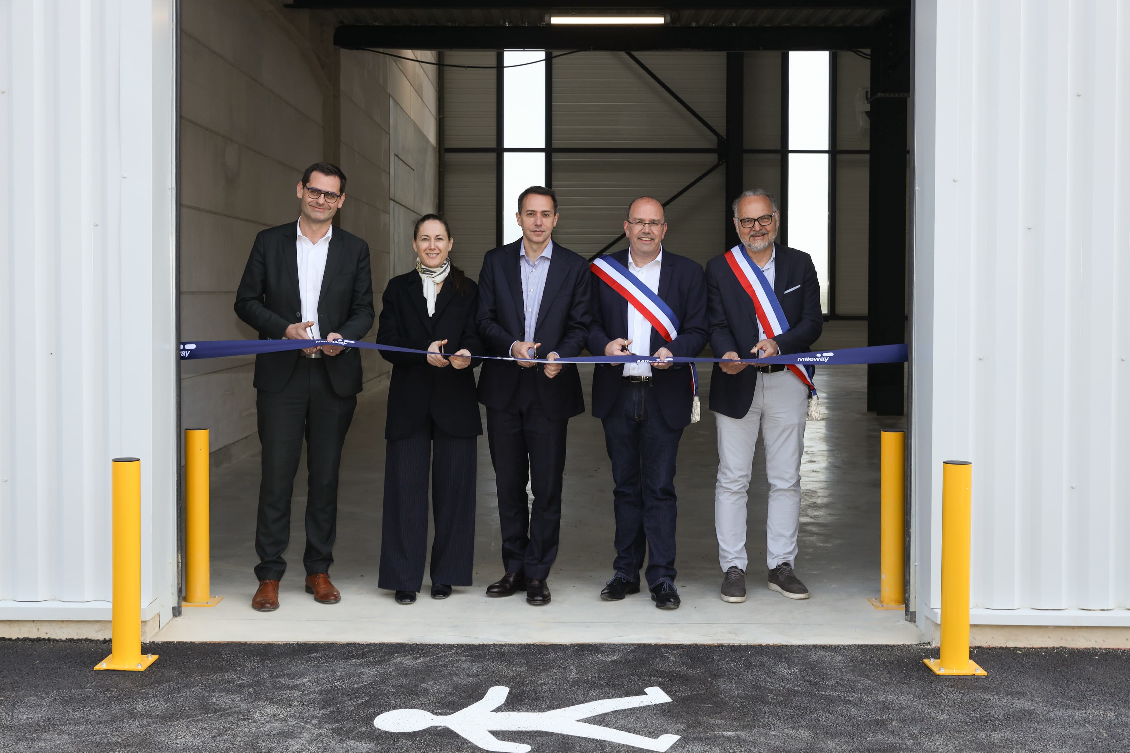 A group in formal attire stands before a building, cutting a blue ribbon at the entrance; two wear tricolour sashes.