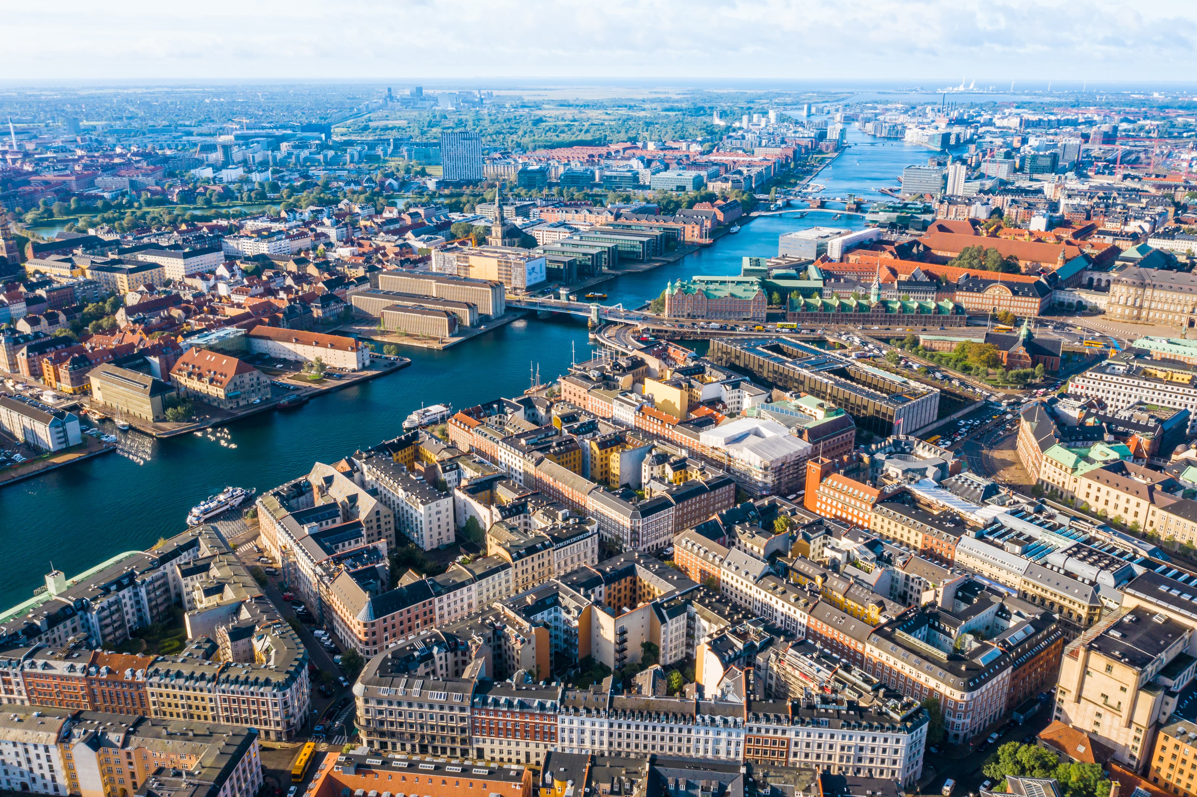 Aerial view of Copenhagen with canals, closely packed buildings, green spaces, and waterways extending far beneath a partly cloudy sky.