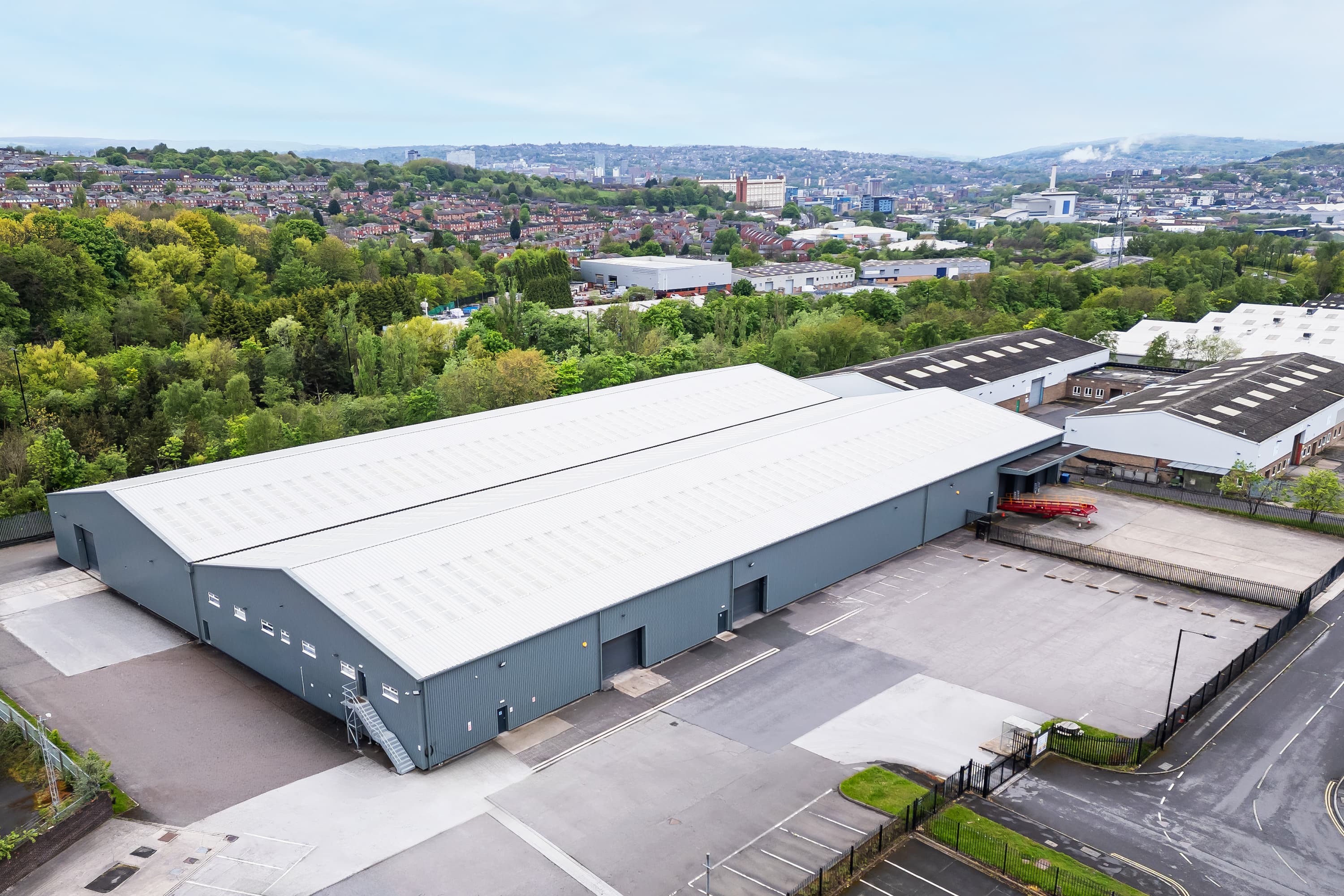 A large grey industrial warehouse with multiple loading bays, paved car parks, and greenery on a city's outskirts with hills behind.
