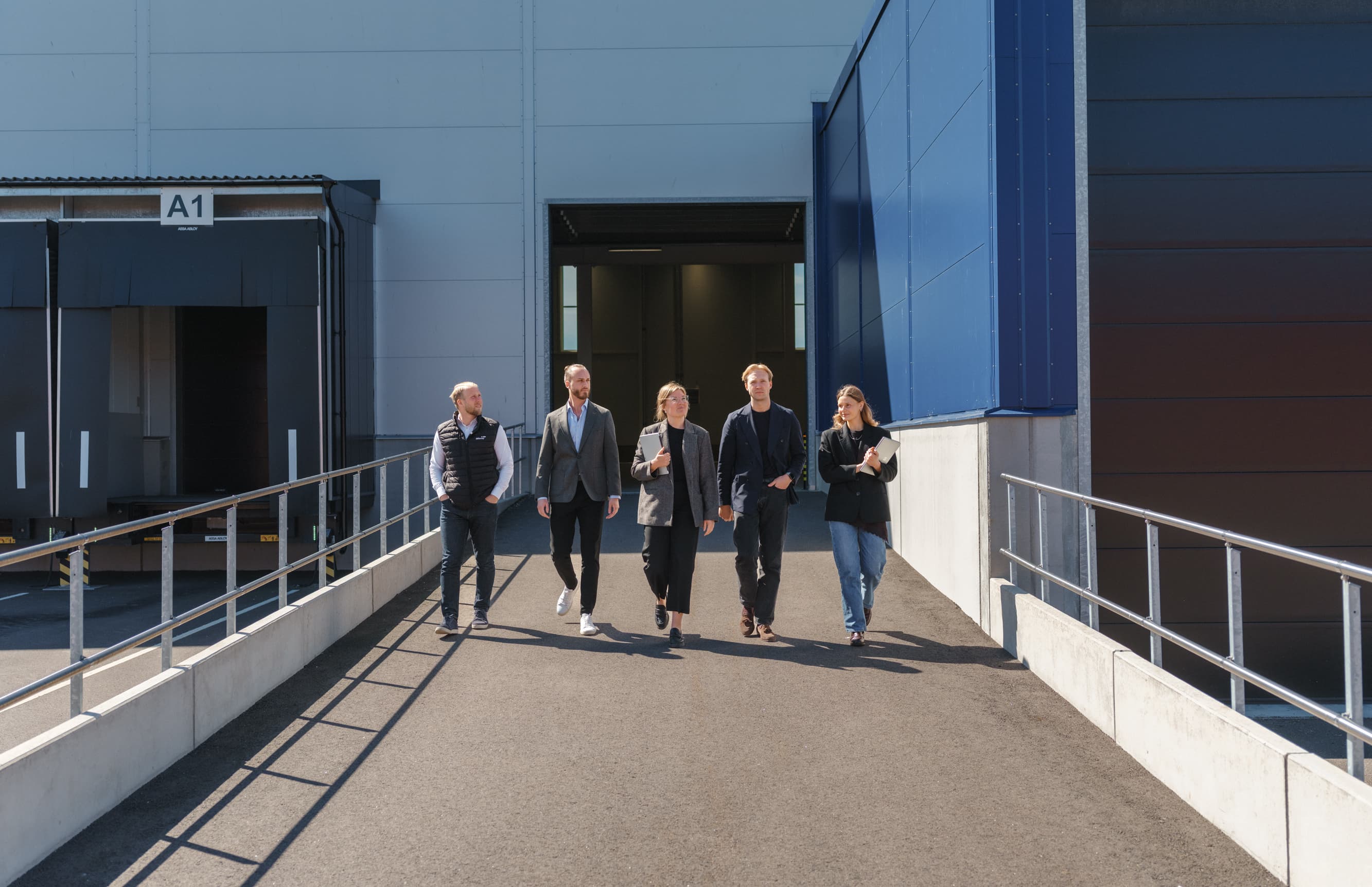 A group in business attire walks down a ramp outside a large industrial building with blue and grey walls.