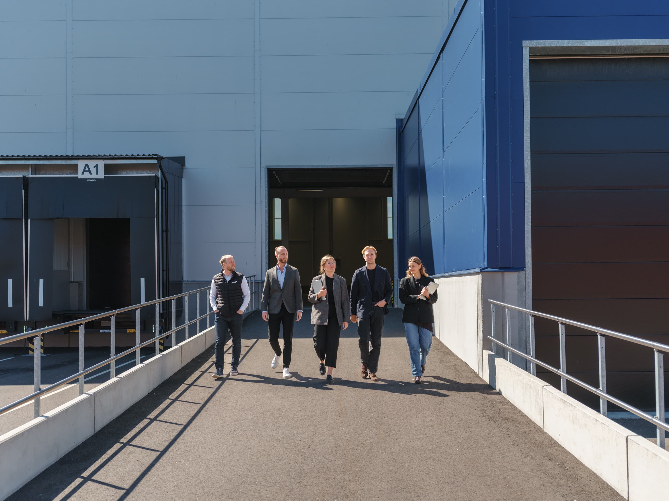 Op een zonnige dag loopt een groep samen een helling af buiten een groot industrie- of magazijngebouw.