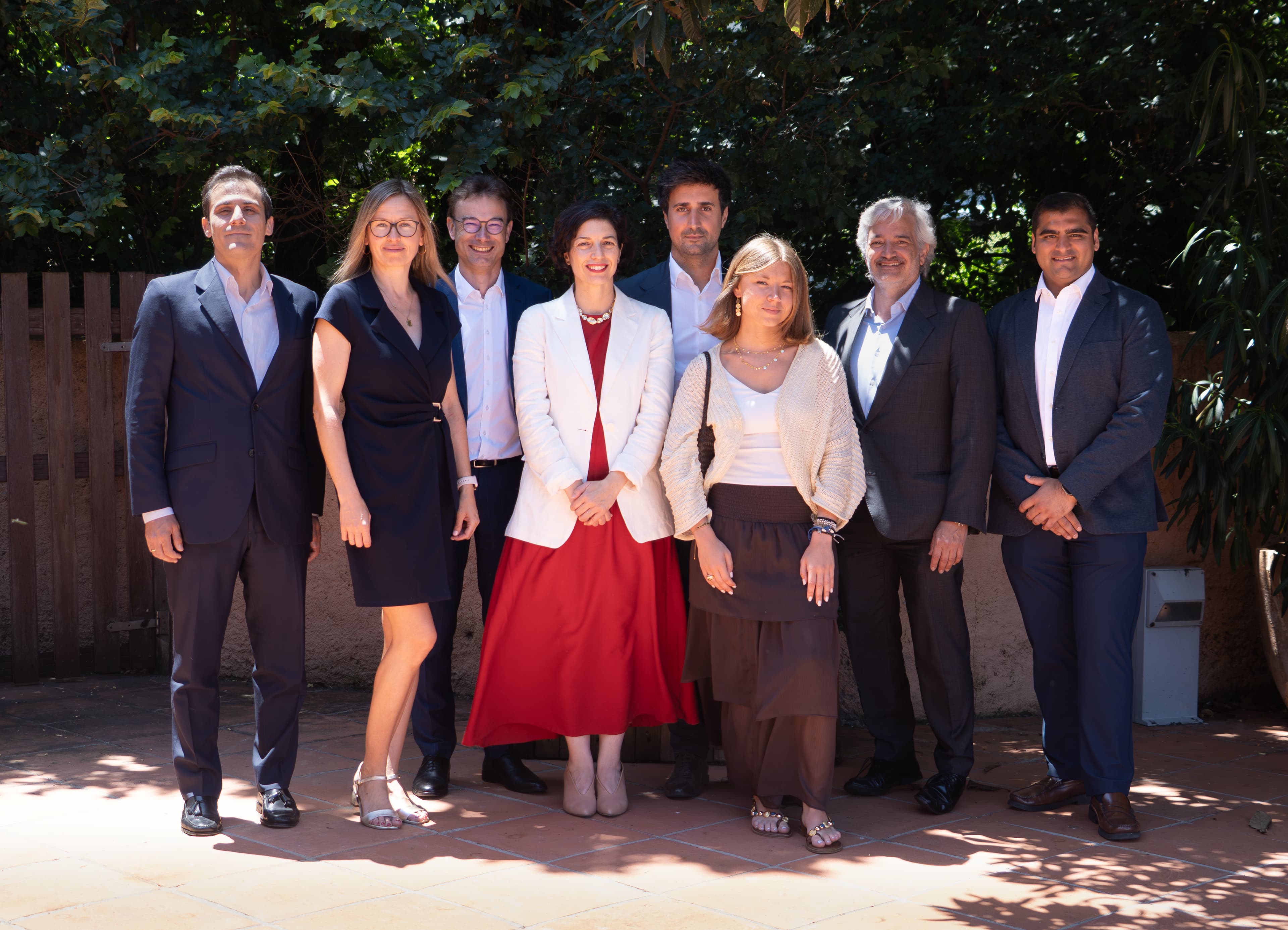 Eight people in business attire stand outdoors on a sunny day, with trees and greenery in the background.