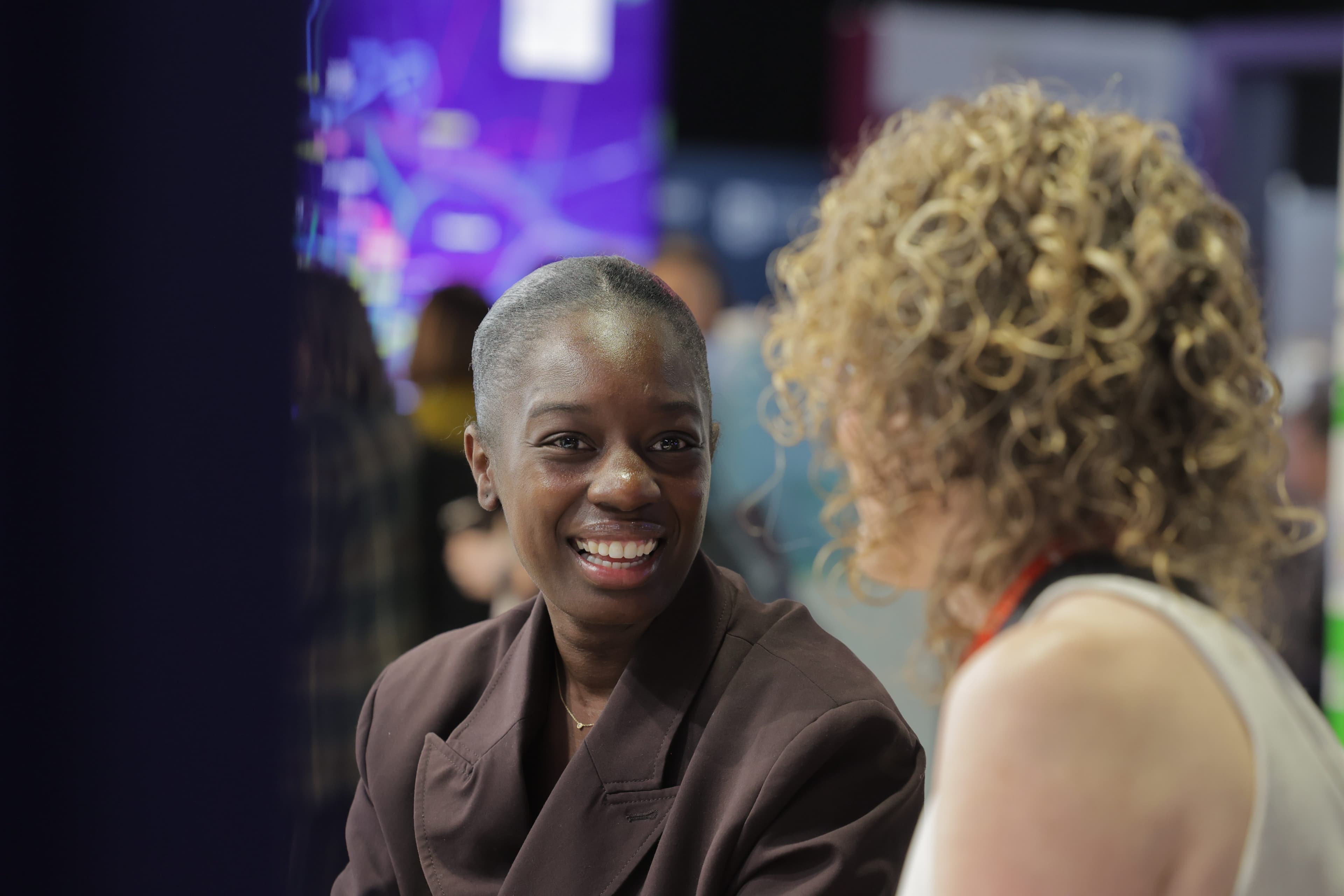 Two people are talking indoors at an event; one is smiling, with a blurred background of colourful lights and others.
