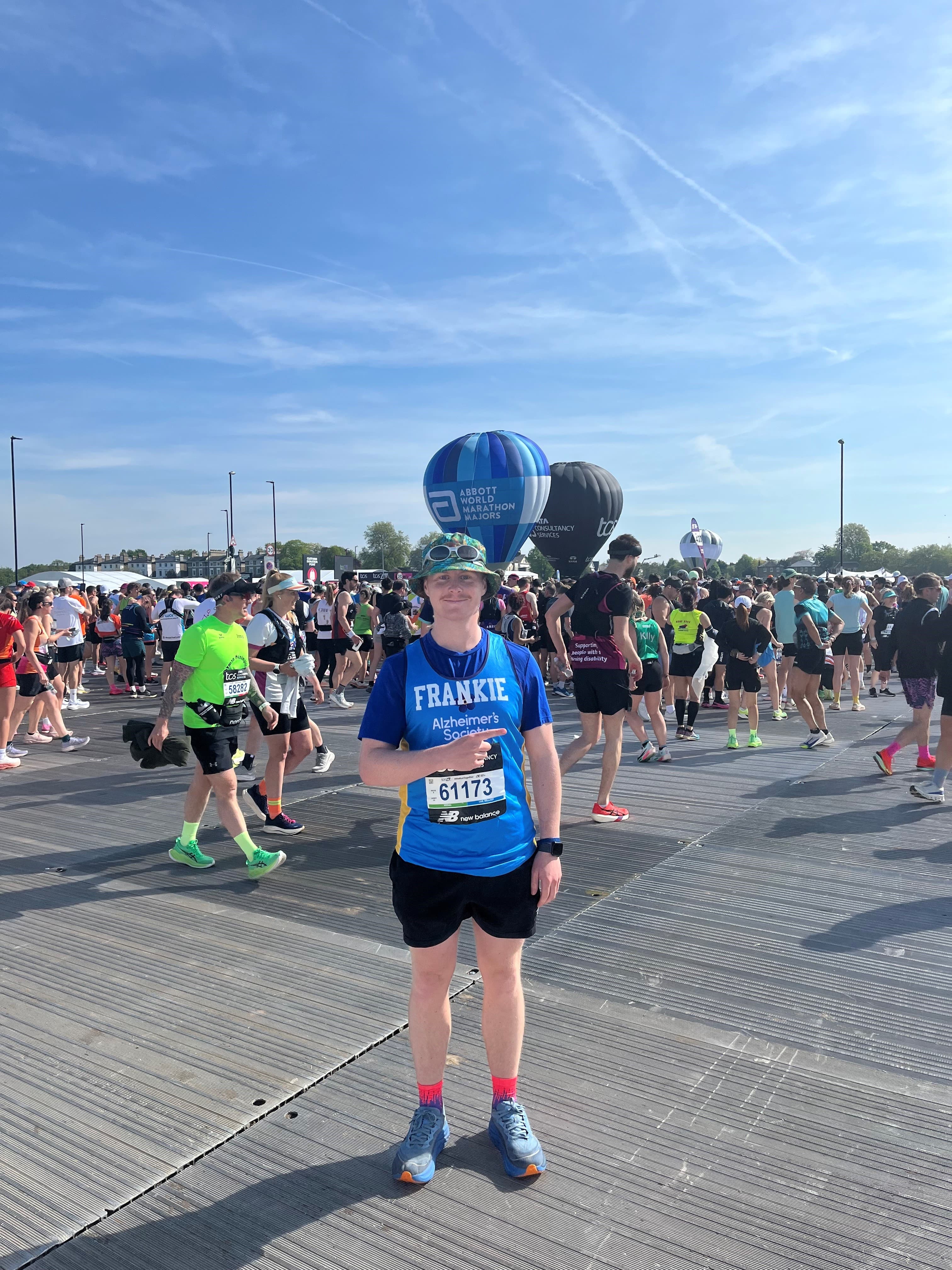 A person in a blue shirt and bib stands before a crowd of runners at an outdoor event, with hot air balloons behind.
