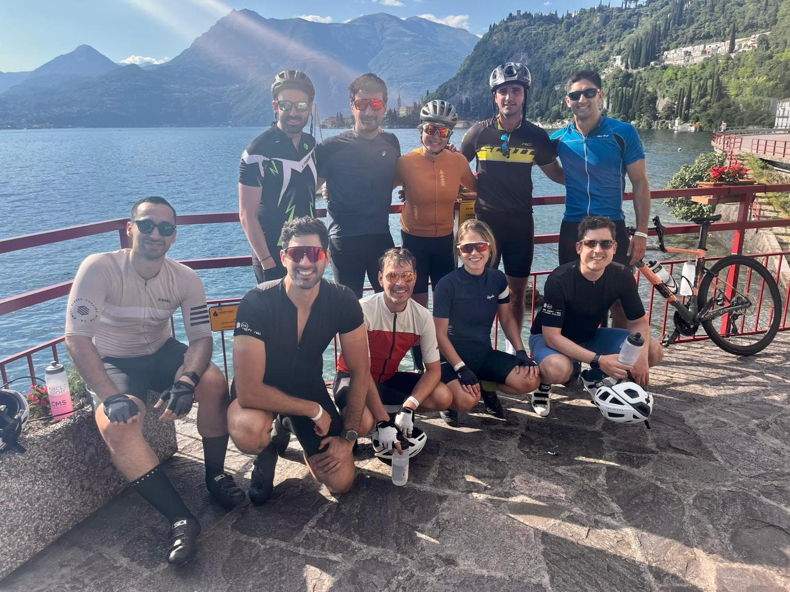 Nine cyclists in helmets and cycling gear pose for a group photo by a scenic lakeside with mountains under a sunny sky.