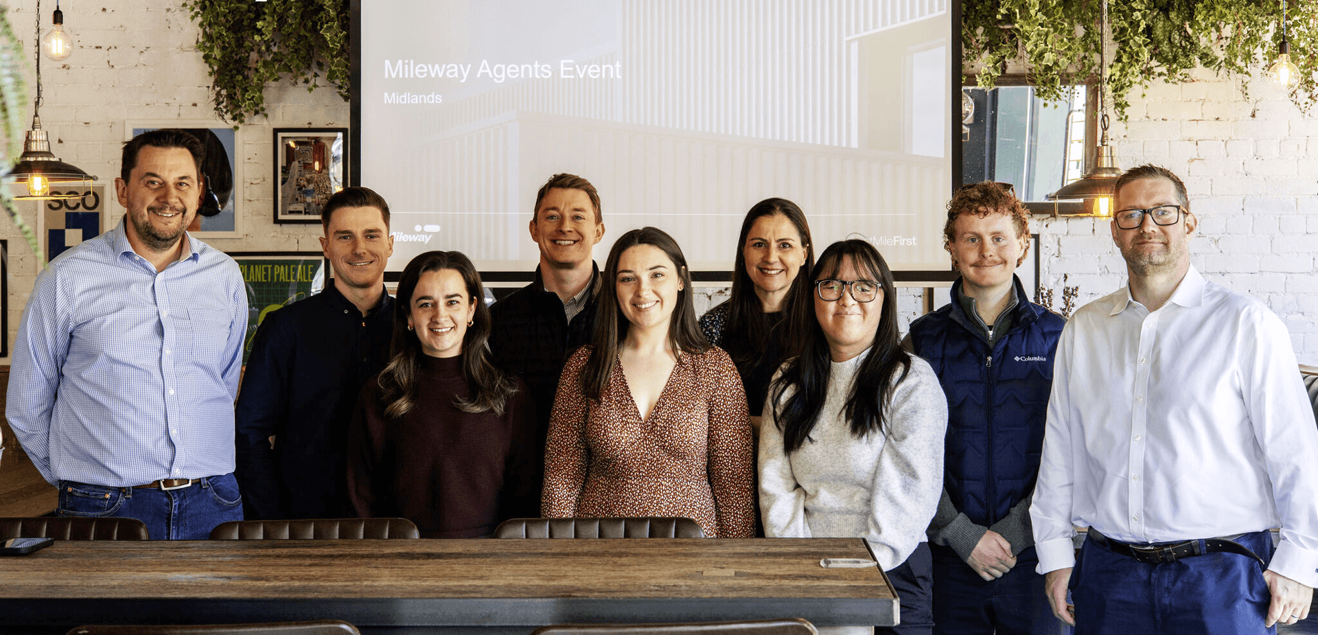 Nine people stand in a row, smiling at the camera in a modern venue.