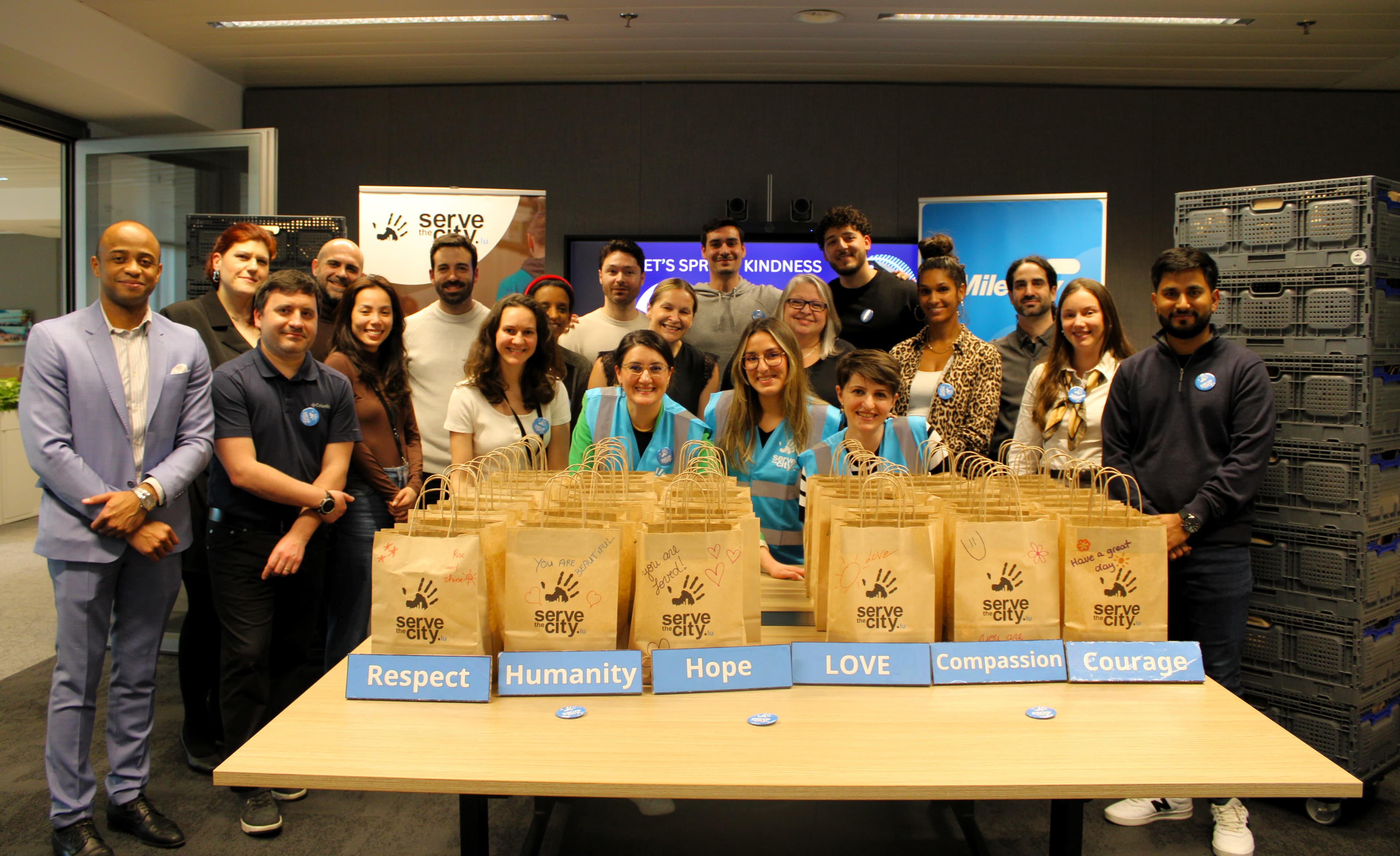 A group stands behind a table with paper bags labelled “serve,” beside signs that say Respect, Humanity, Hope, Love, Compassion, and Courage.