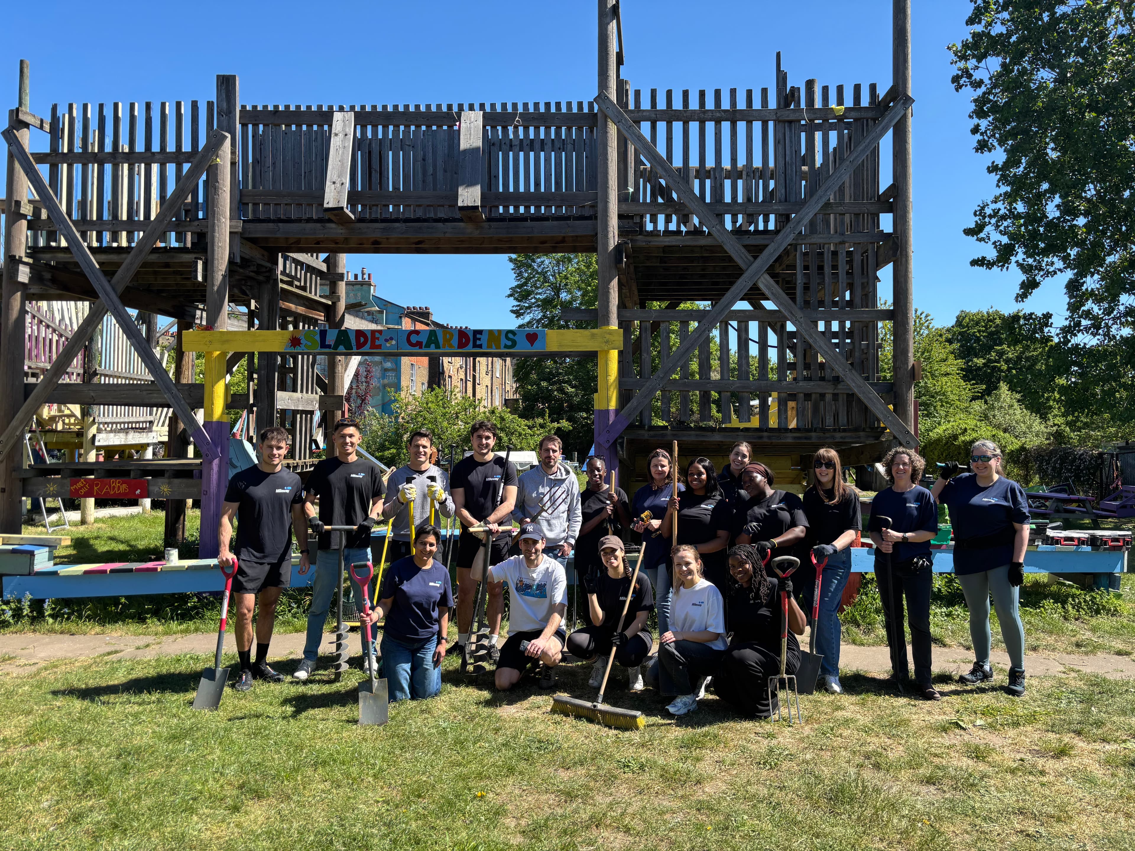 People stand with spades in front of a wooden playground structure under a LADE GARDENS sign on a sunny day.