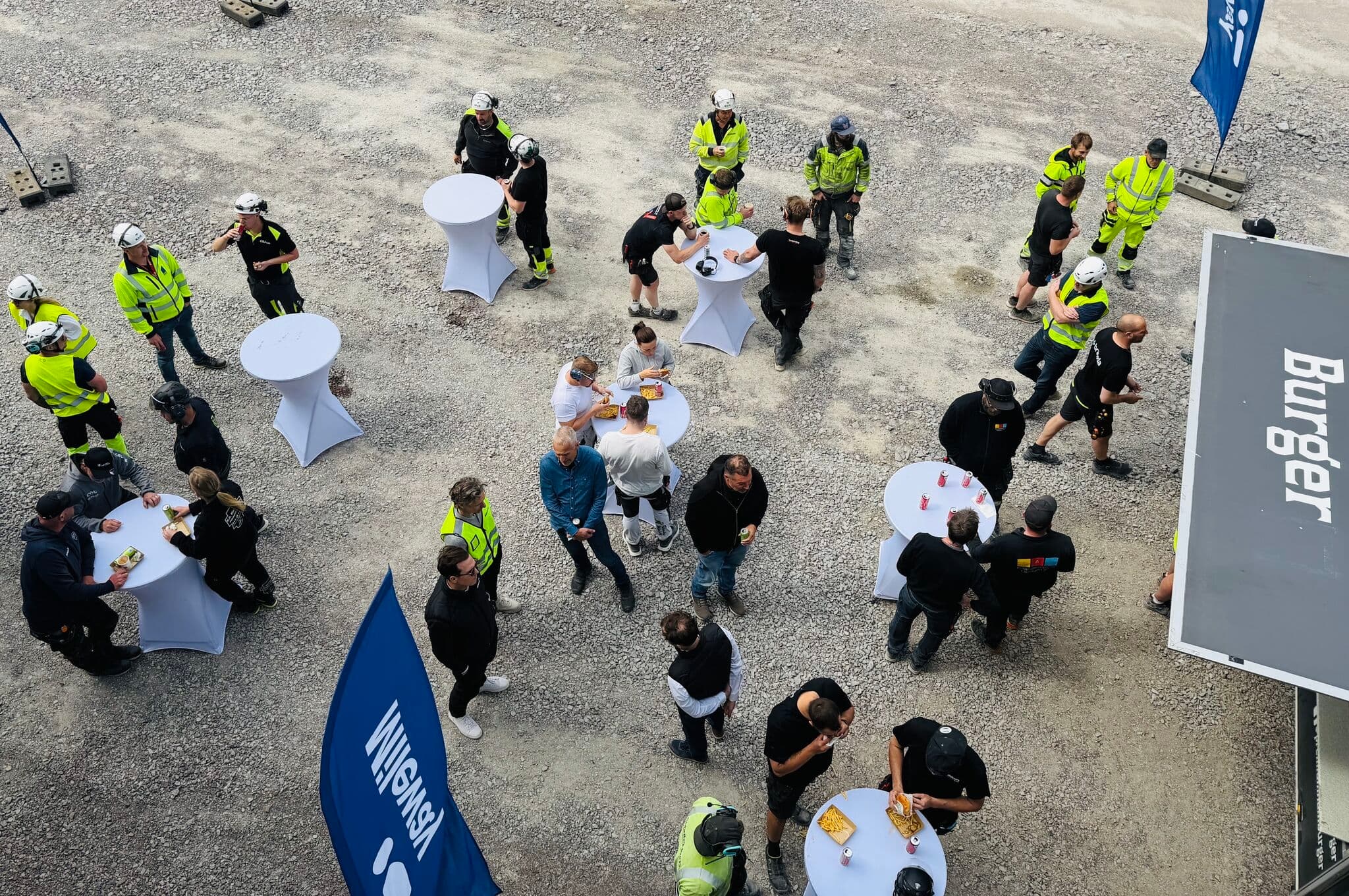 People in safety gear and casual clothing gather at outdoor cocktail tables on gravel, eating near a food van and blue banners.