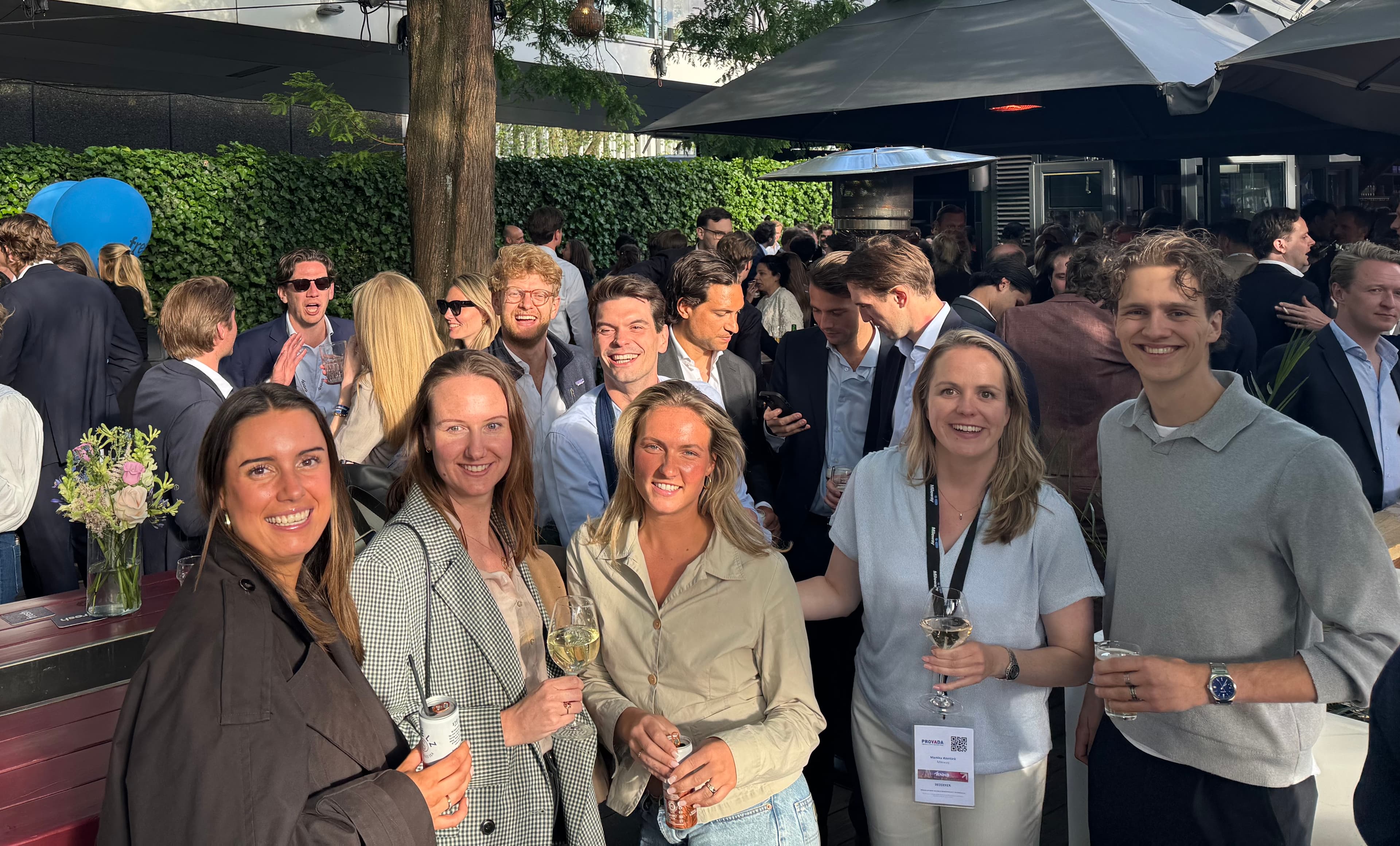 A group at an outdoor social event, some holding drinks, with others mingling in the background on a sunny day.