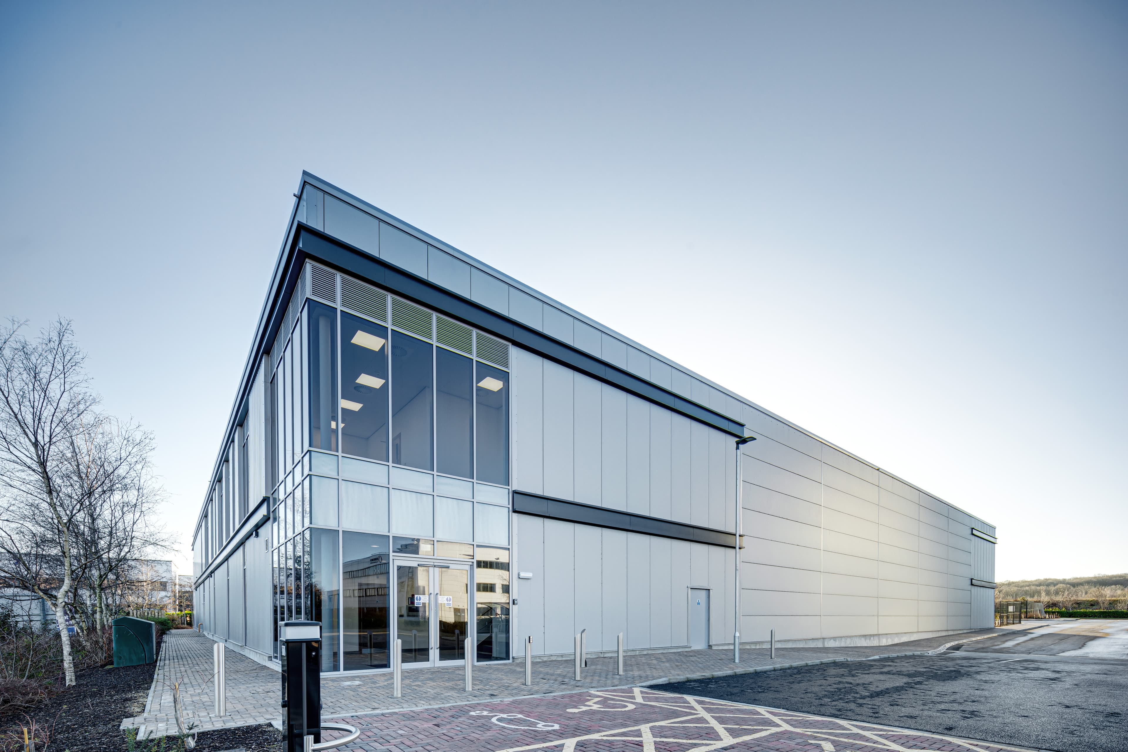 A modern industrial building with glass windows and metal cladding beside a tarmacked road and marked parking spaces on a clear day.
