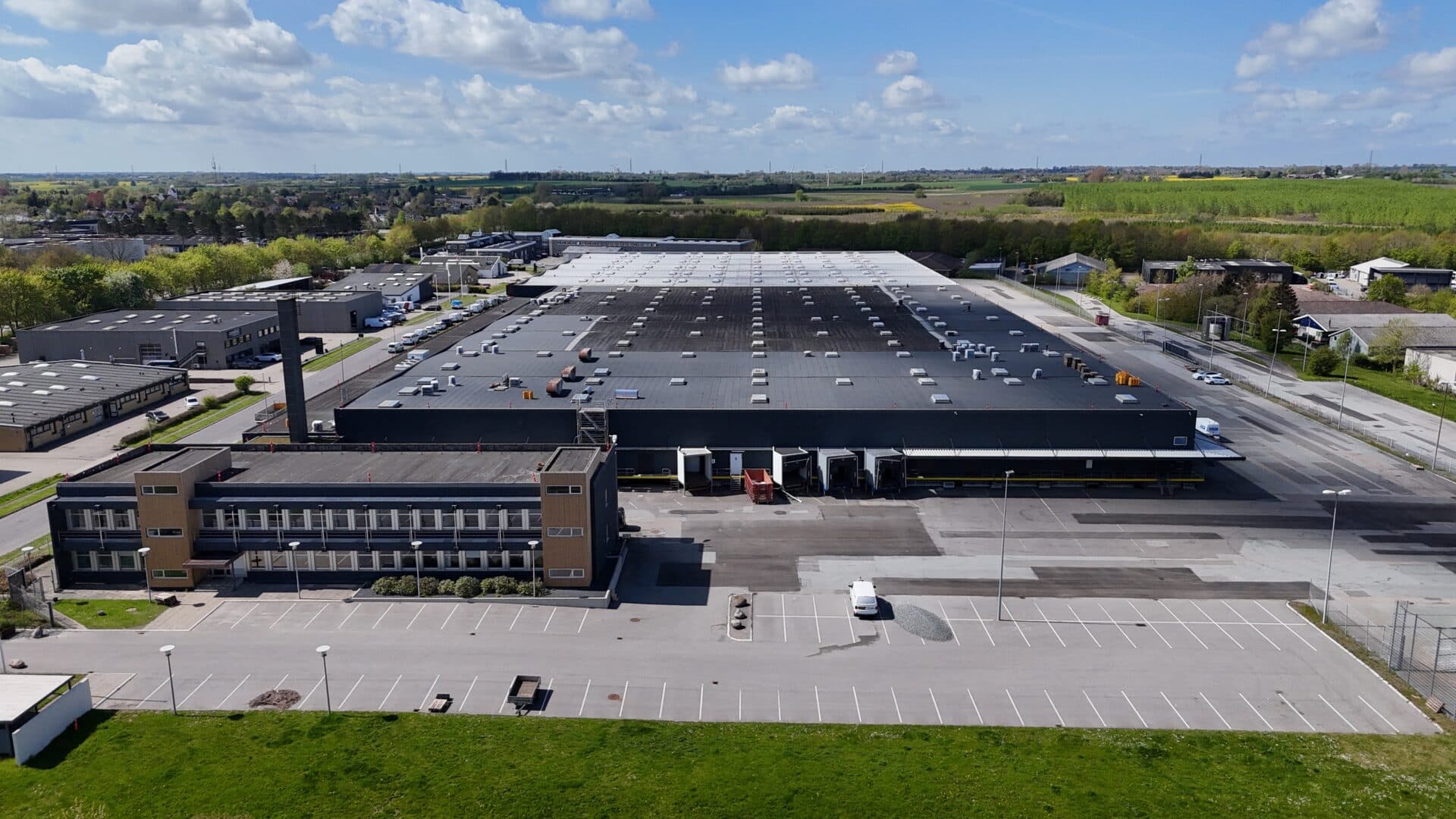 A large industrial warehouse with a dark roof and a smaller office building, surrounded by car parks and green fields under a cloudy sky.