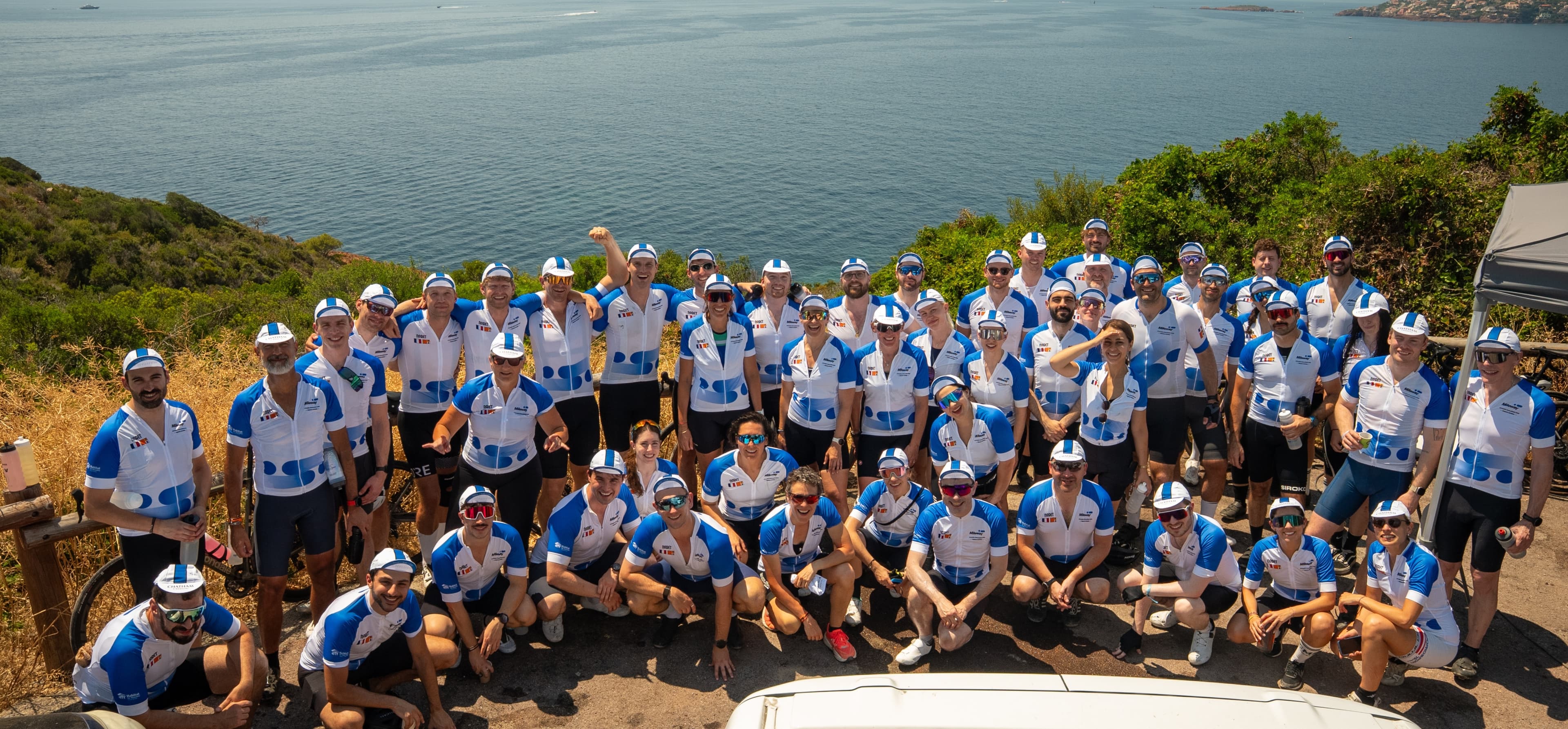 A large group of cyclists in matching blue and white Mileway jerseys and caps pose outdoors near the sea with coastal scenery behind them.