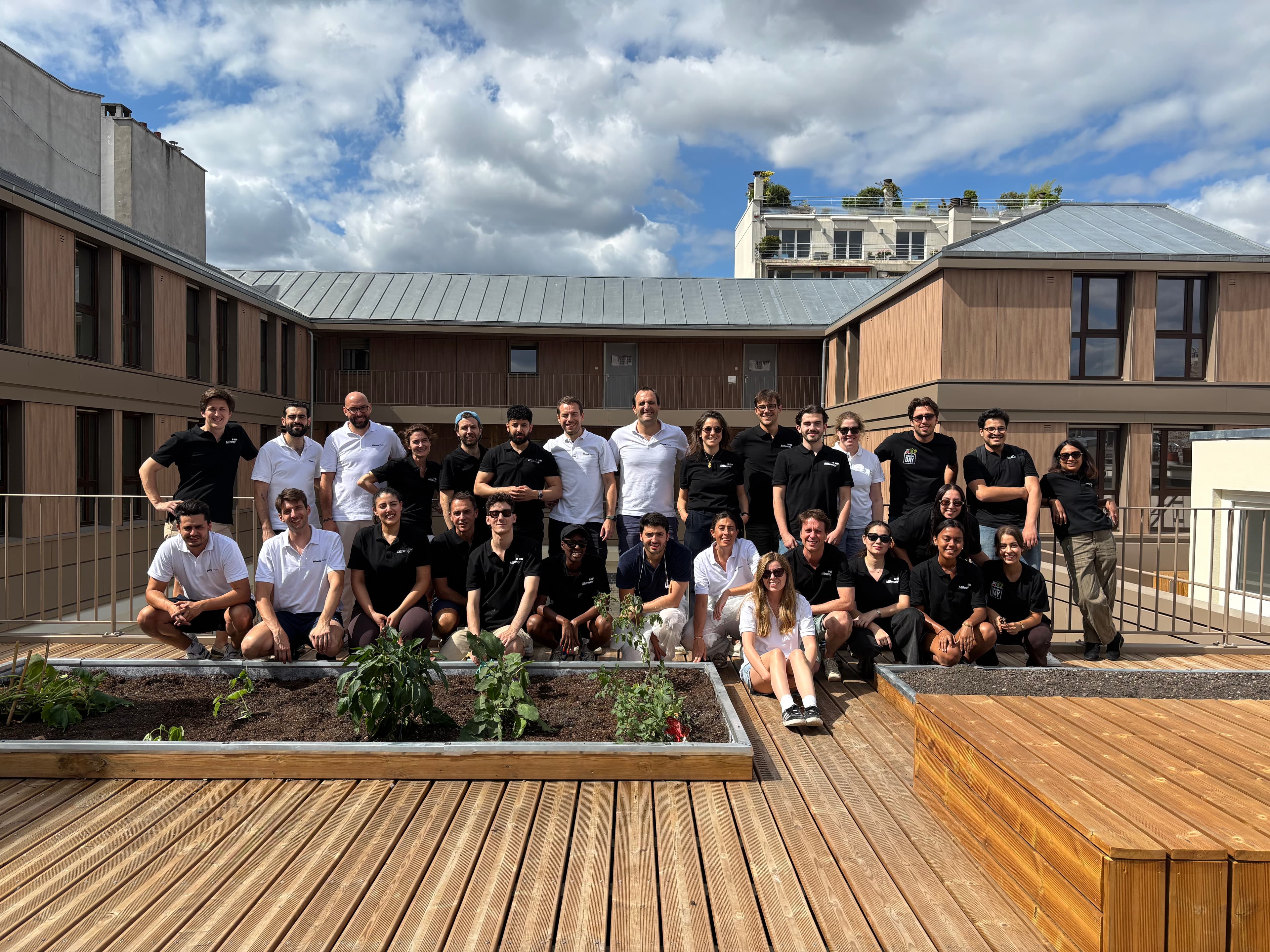 A group poses for a photo on a wooden rooftop terrace with planters, modern buildings, and a cloudy sky in the background.