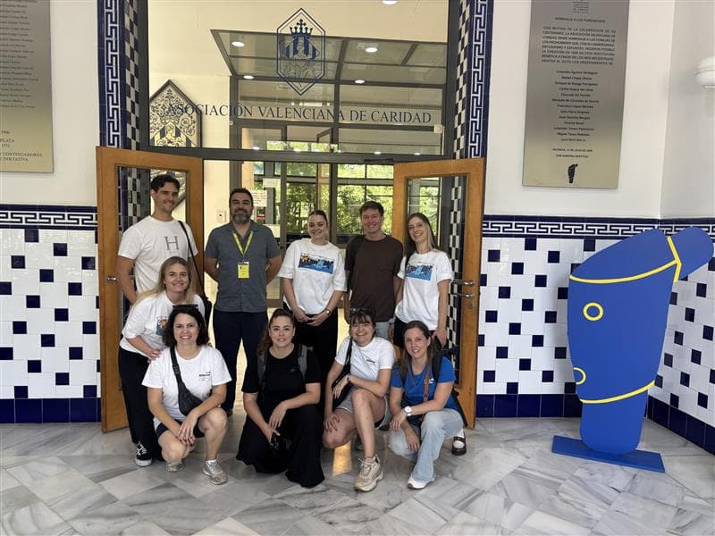 A group of ten people pose for a photo inside a building with blue and white tiled walls and a sign reading “Asociación Valenciana de Caridad” above the entrance.