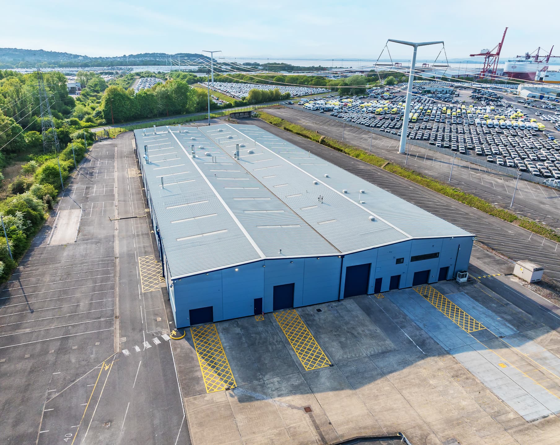 A large blue warehouse sits in an industrial area with empty parking spaces, surrounded by trees, parked cars, and cranes in the background under a clear sky.