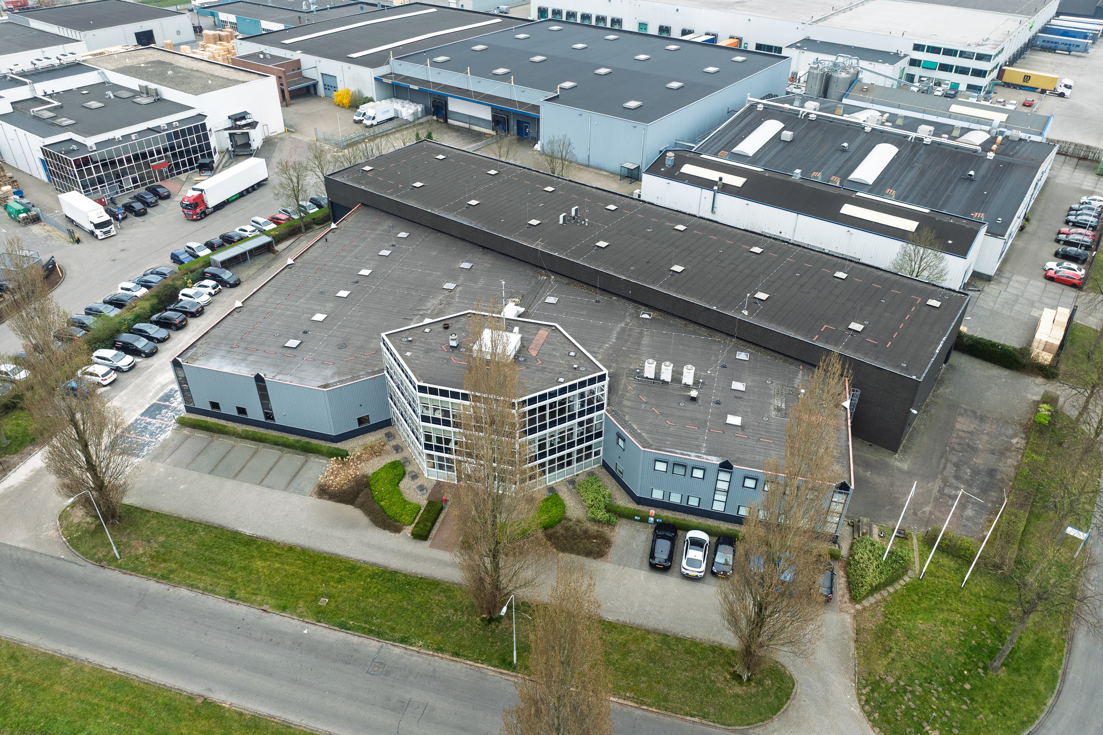 Aerial view of an industrial building with a car park, surrounded by trees and other warehouses in a business park.