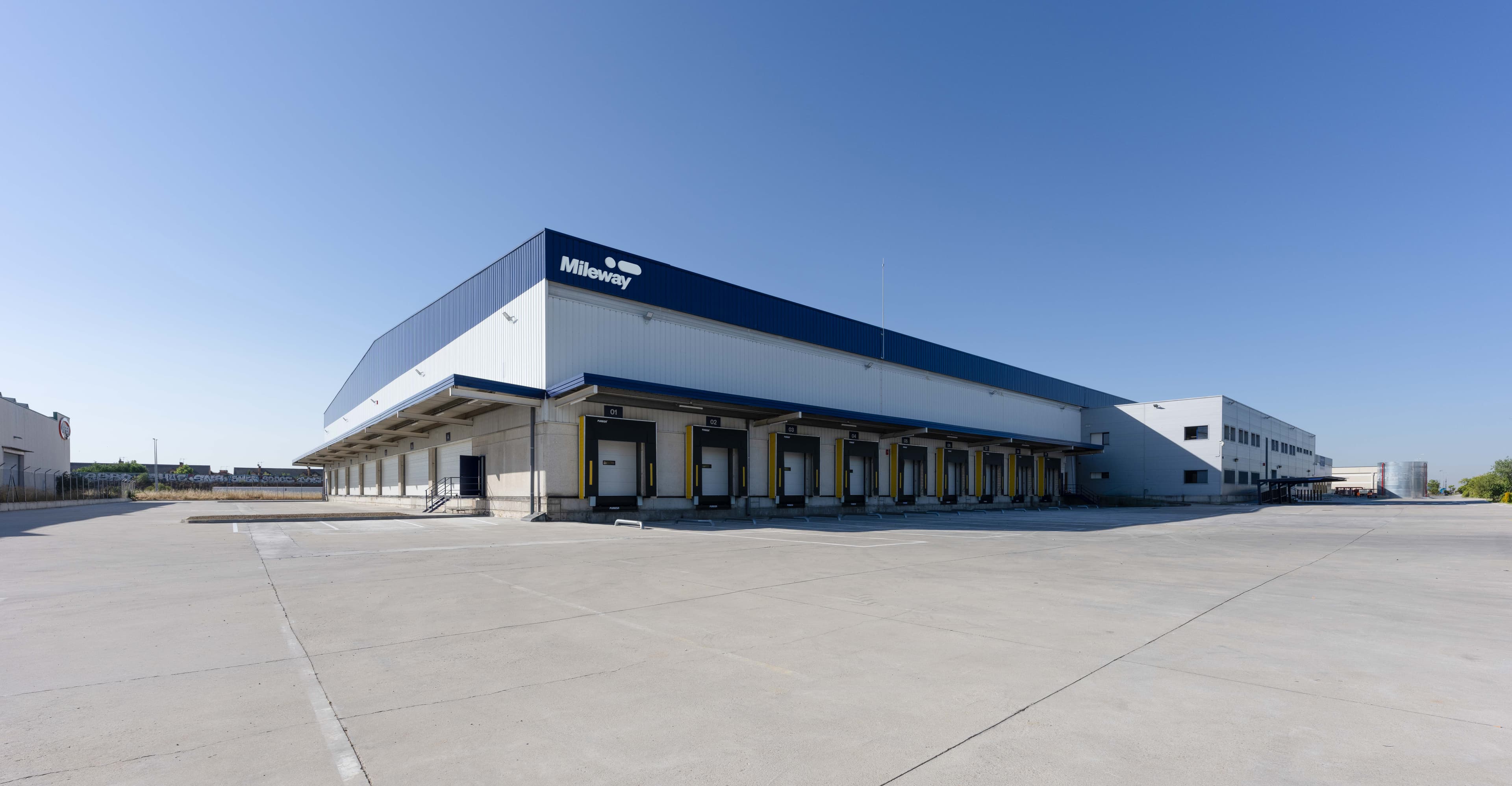 Large industrial warehouse with multiple loading bays, featuring a blue and white exterior and spacious empty concrete area in front, beneath a clear blue sky.