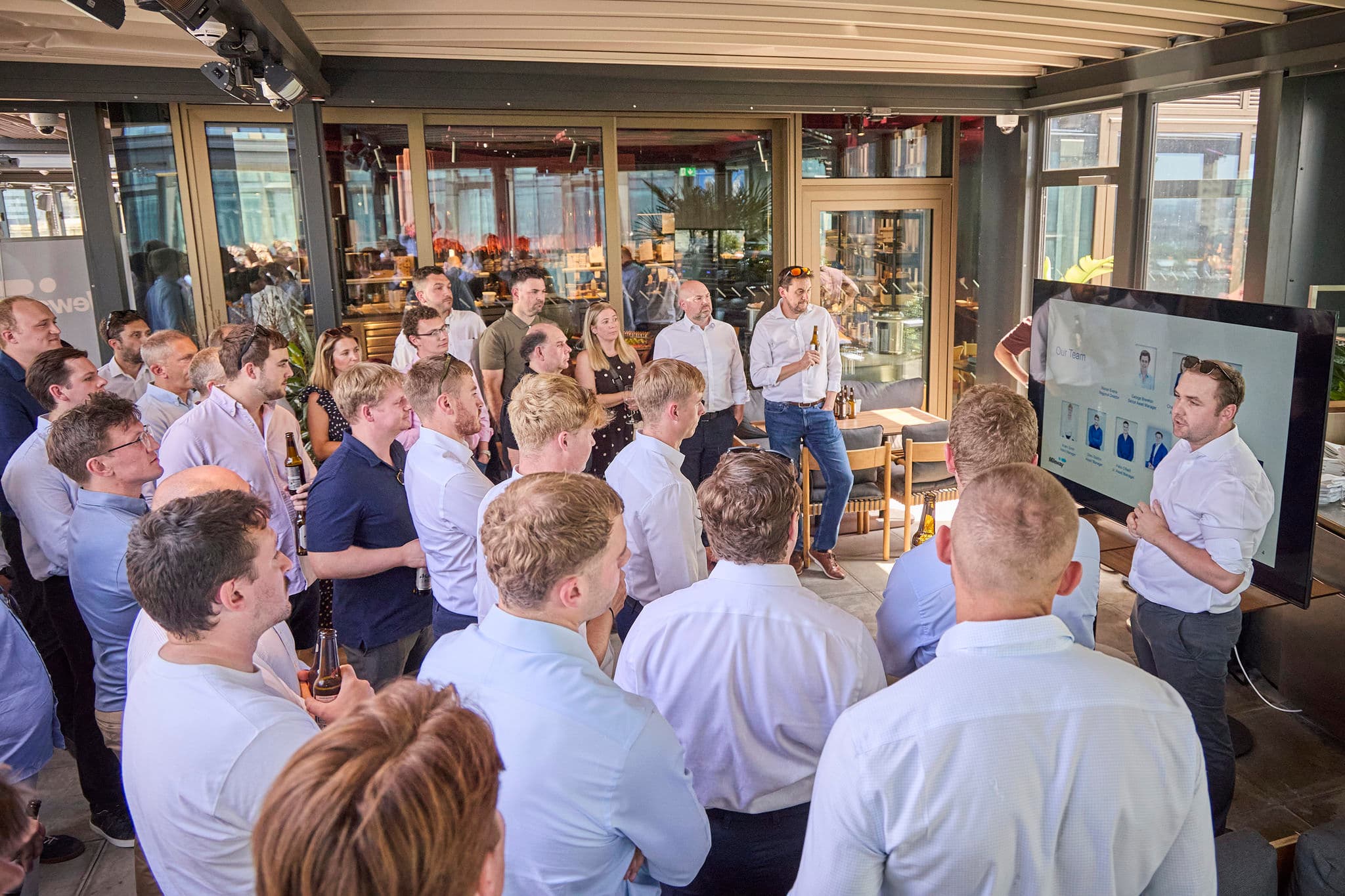 A group of people in business attire listen to a presenter sharing slides on a large screen inside a modern, glass-walled venue.