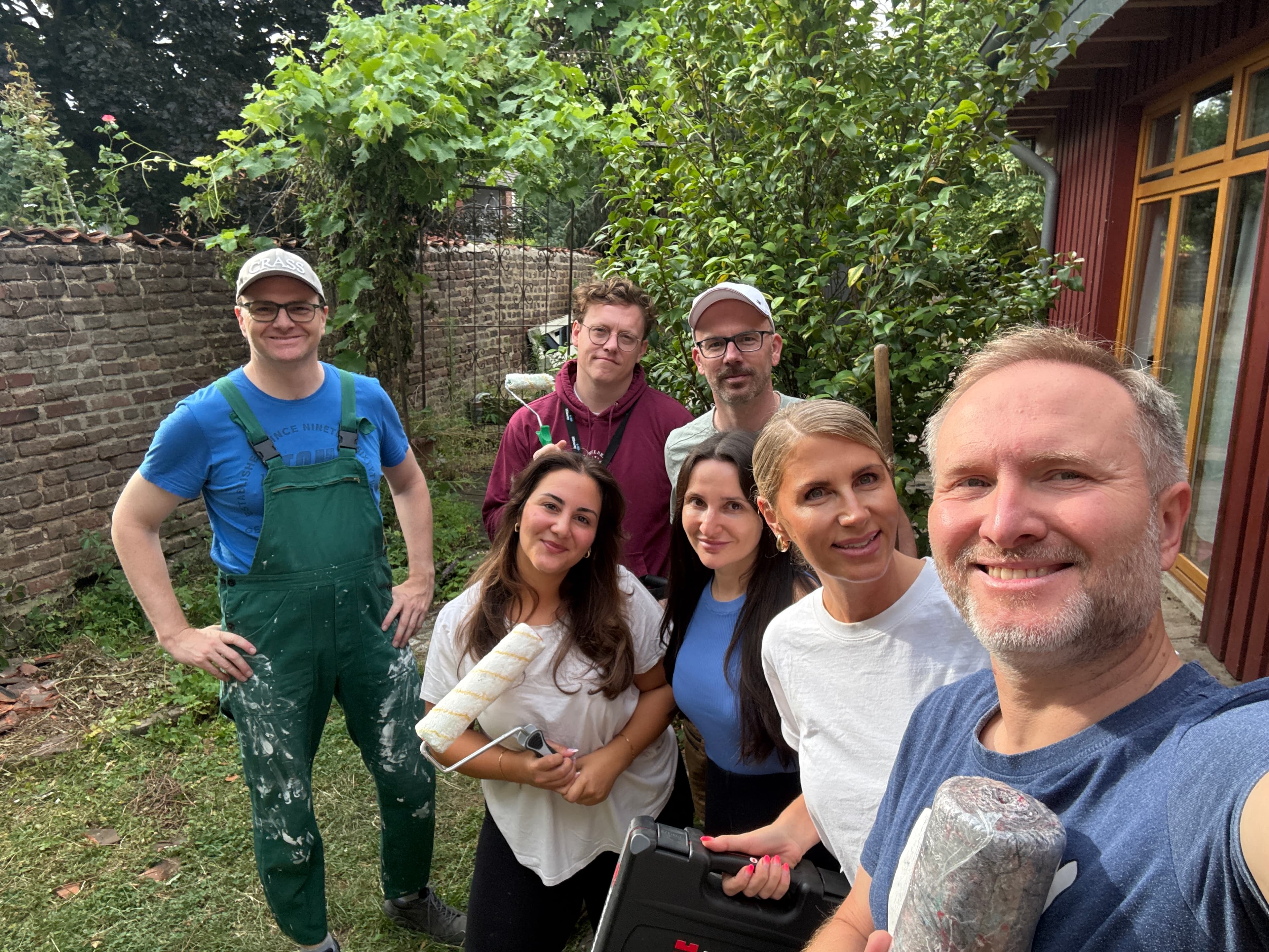 Seven people posing in a garden, some holding painting and renovation tools, with greenery and a brick wall in the background.