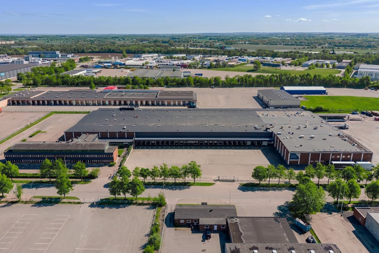 Aerial view of a large industrial warehouse complex with multiple loading bays and surrounding car parks, situated in a semi-rural setting with trees and open land in the background.