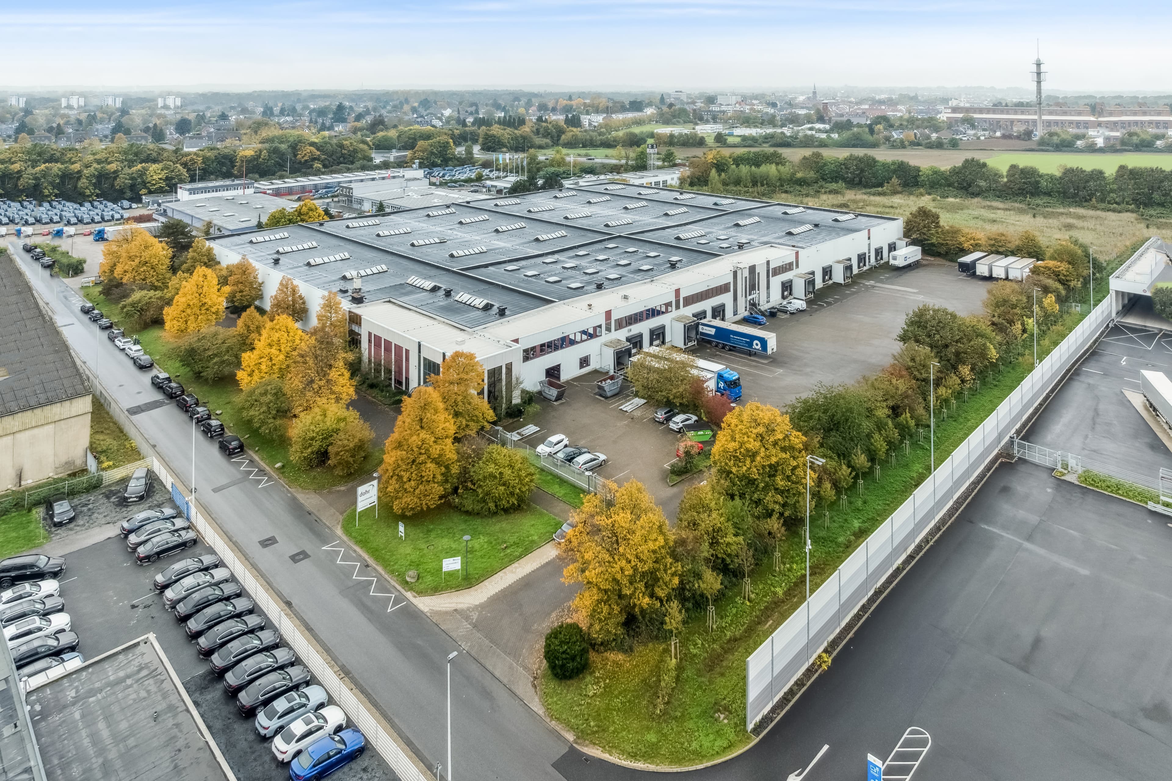 A large industrial warehouse with solar panels on the roof, surrounded by trees, car parks, and a fence, with lorries and cars parked outside.