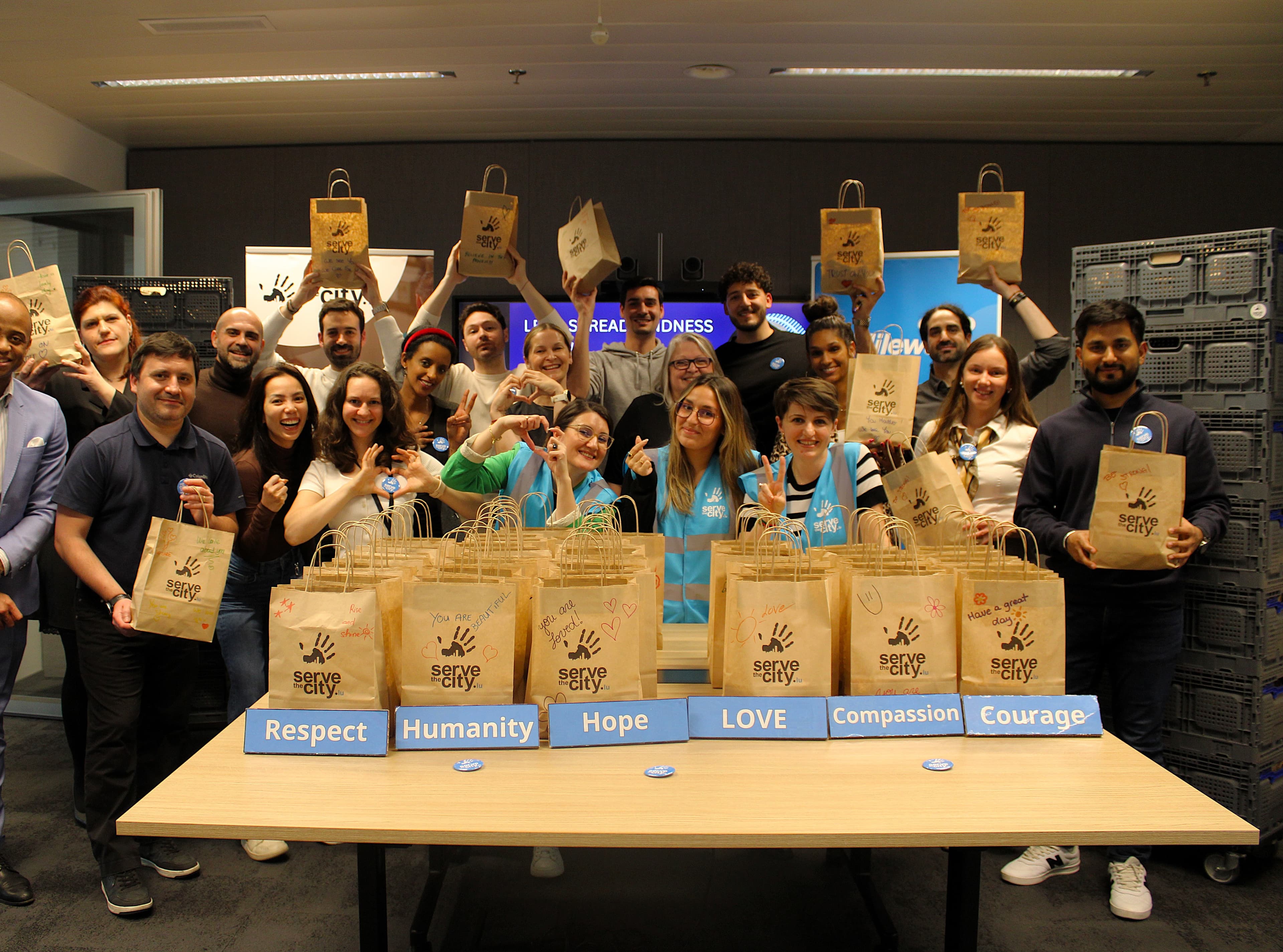 A group of people pose indoors behind a table with gift bags and signs reading Respect, Humanity, Hope, LOVE, Compassion, and Courage. Most are smiling and holding up bags.
