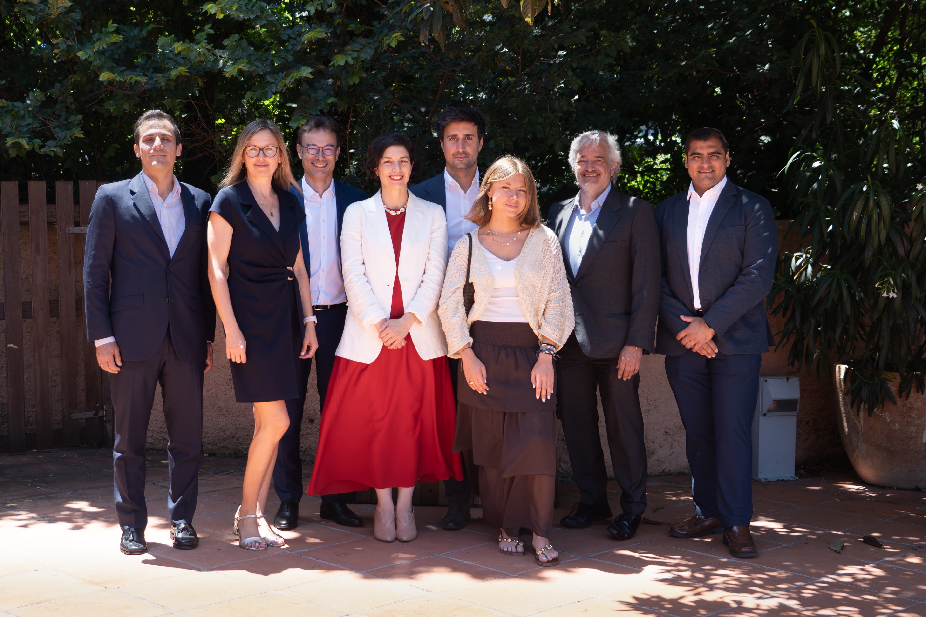 Un grupo de ocho personas está de pie en fila al aire libre, delante de un árbol y una valla de madera, sonriendo para una foto de grupo.