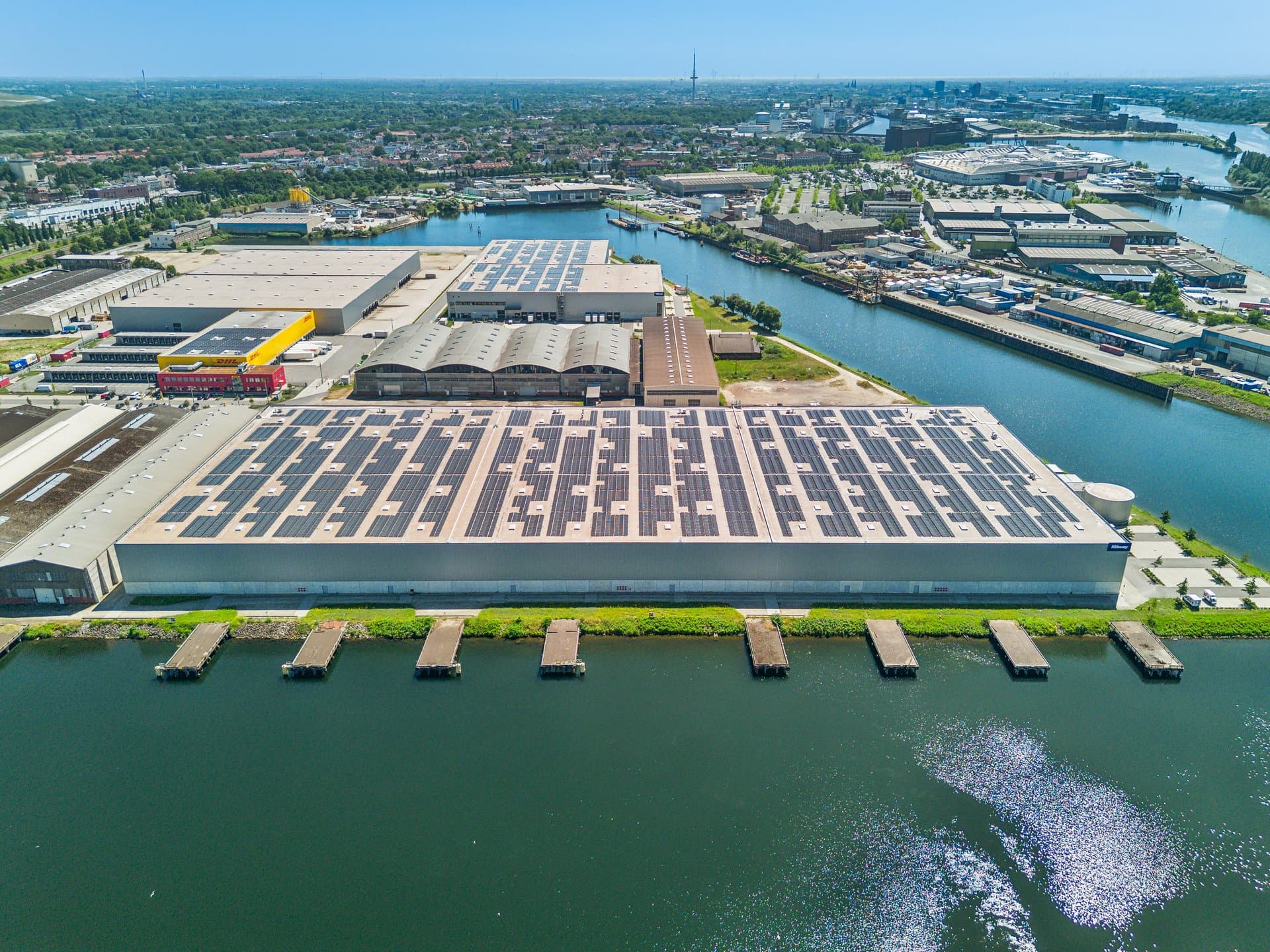 Aerial view of a large warehouse with solar panels on the roof, situated by a waterway in an industrial area among other buildings and docks.