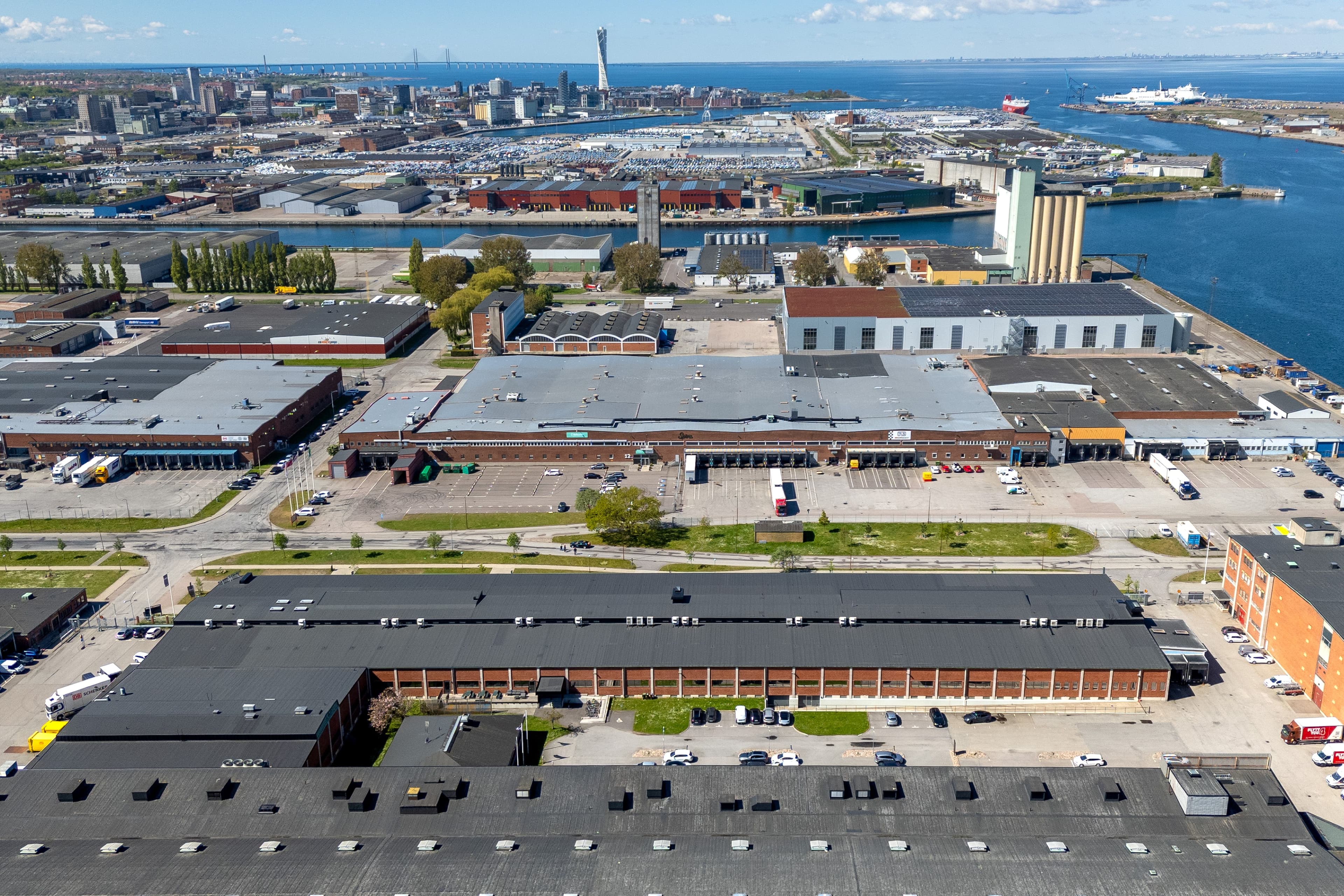 Aerial view of industrial buildings and warehouses near a waterfront, with a city skyline and ships in the background under a partly cloudy sky.