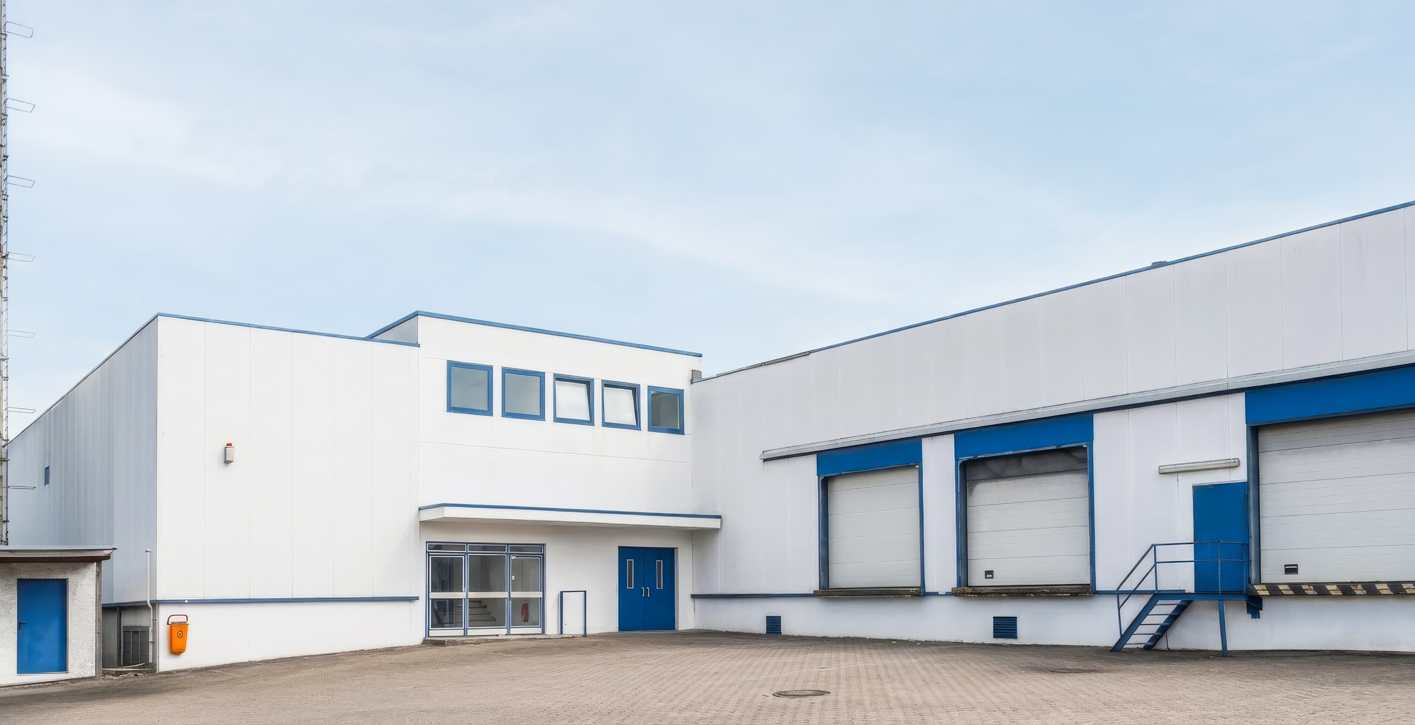 A modern warehouse with white walls, blue trim, multiple loading bays, and an entrance under a clear sky.