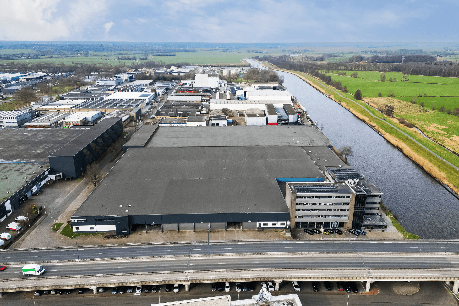 Aerial view of an industrial area with large warehouses, an office block, a road, and a canal running beside grassy fields.