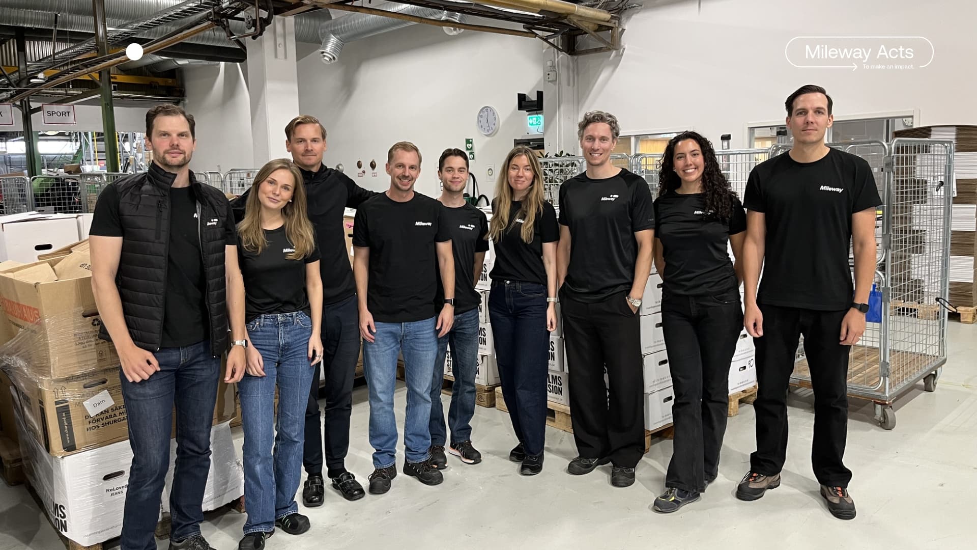 Ten people stand in a warehouse-style space, dressed in matching black shirts and jeans or black trousers, posing in front of stacks of boxes and metal trolleys.