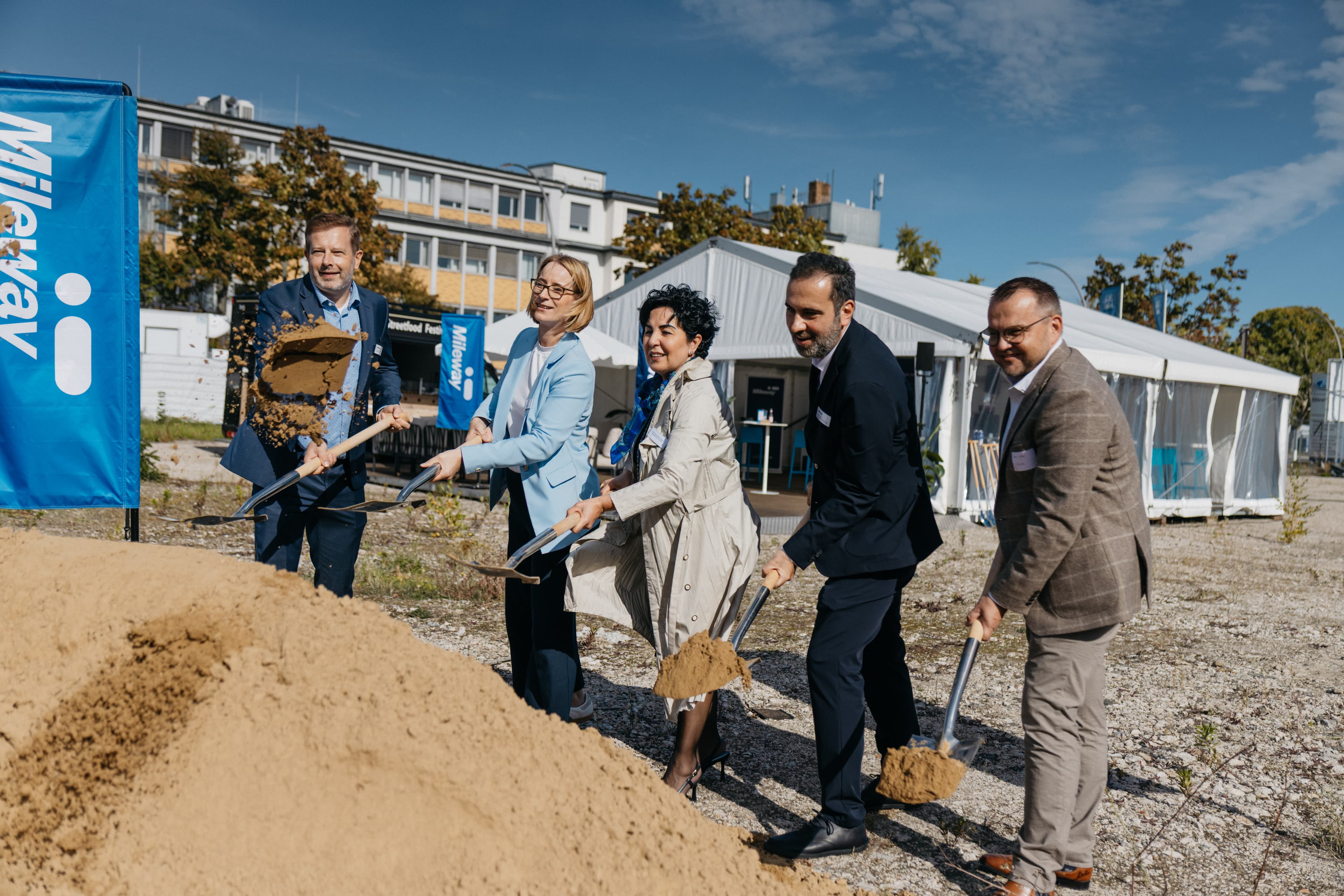 A group in business attire uses spades to move soil at an outdoor groundbreaking ceremony, with a marquee and building in the background.
