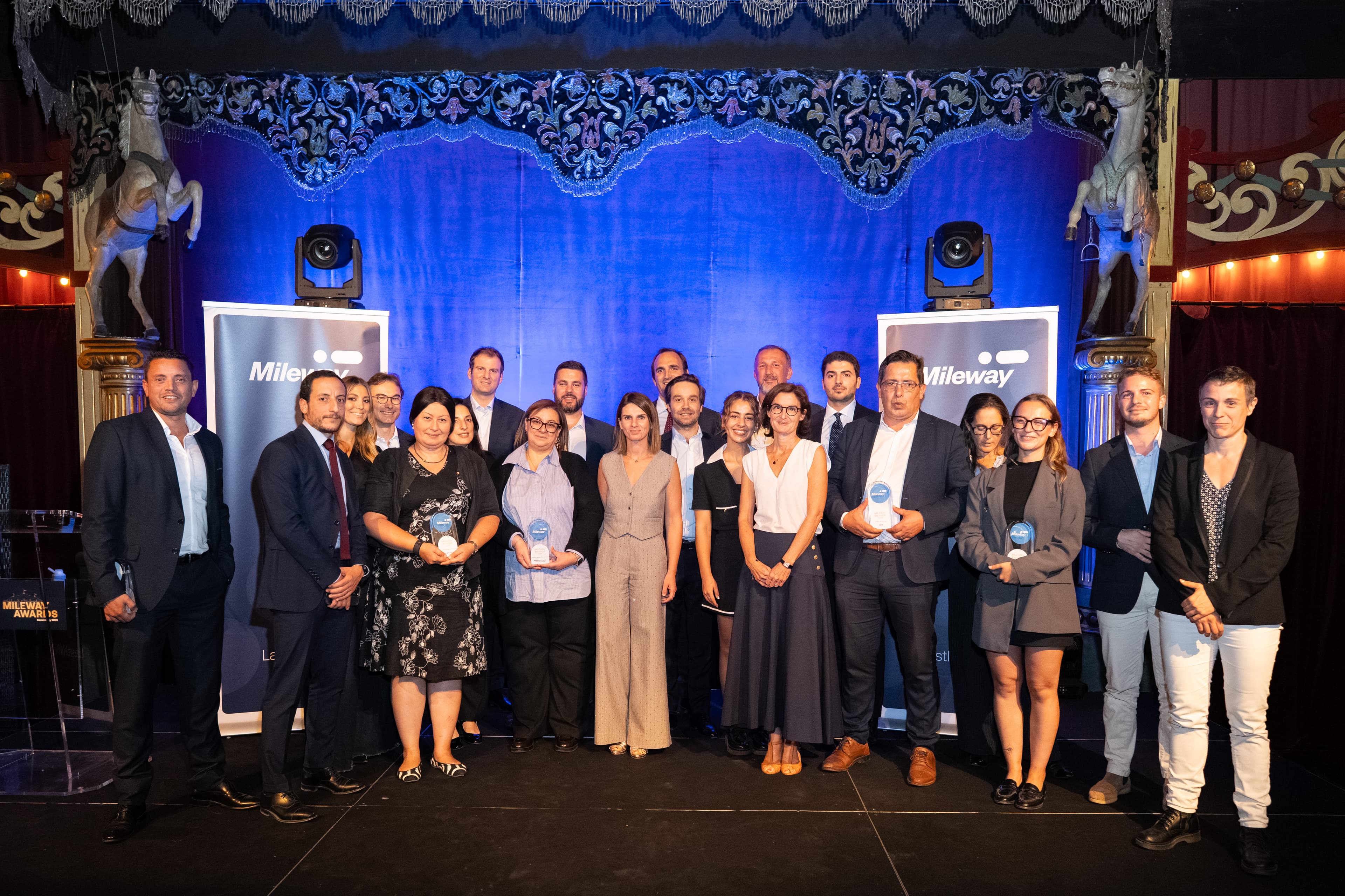 A group of people standing on a stage in front of a blue backdrop with Mileway banners and two awards.